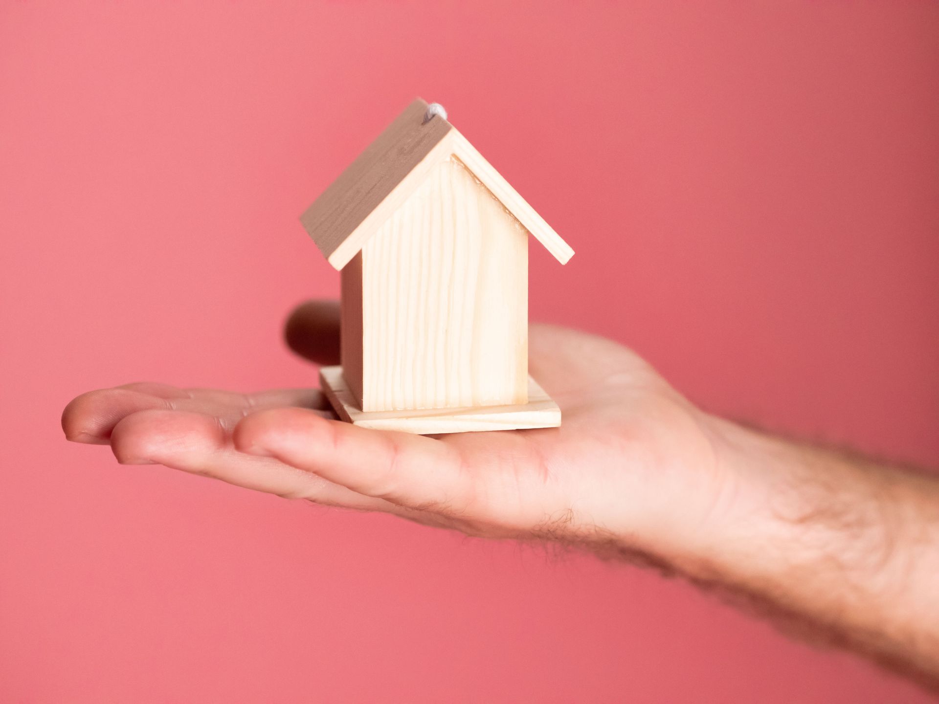A hand holding a small wooden house model against a pink background.