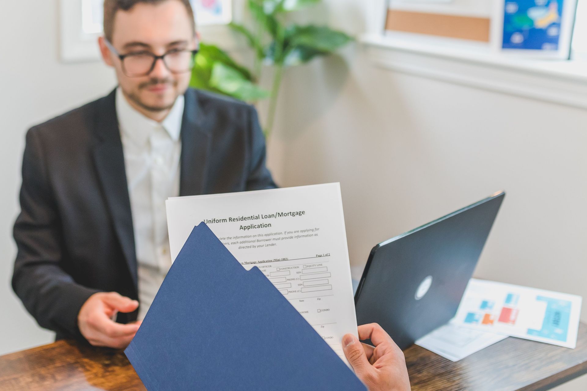 Man reviewing paperwork across a desk with a laptop and blue folder in a bright office
