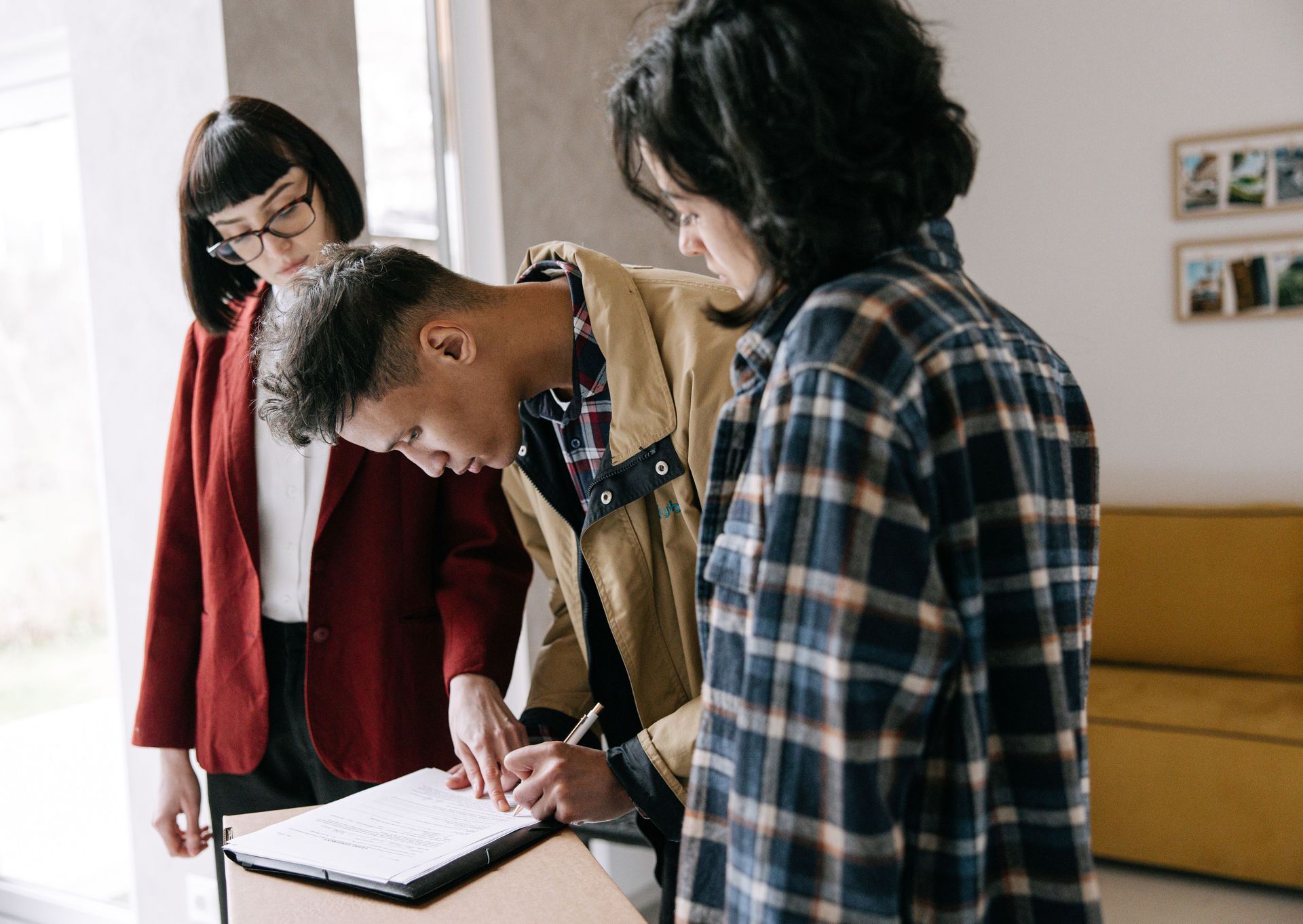 Three people lean over a notebook at a desk, discussing work in a bright office.