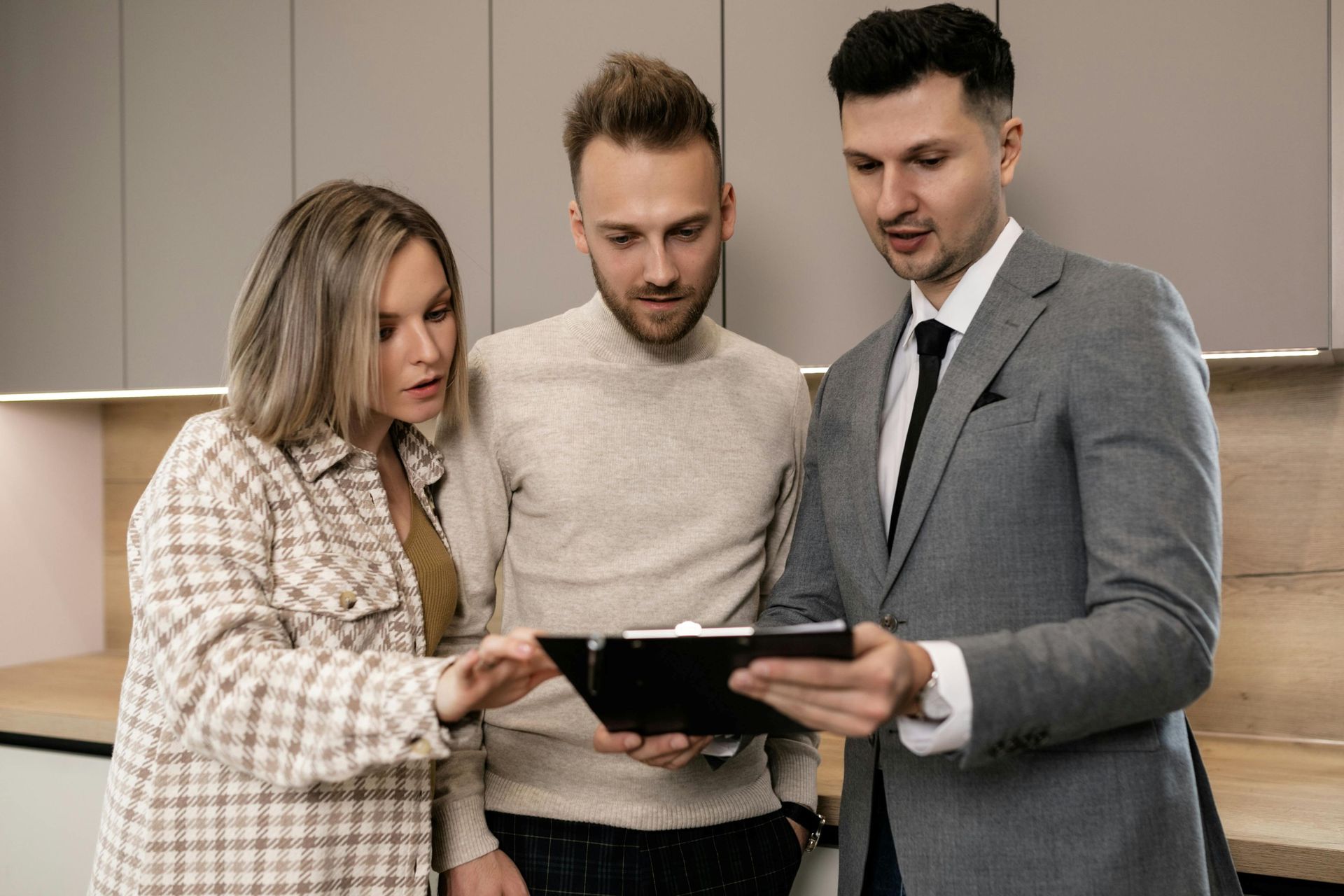 Three people look at documents on a clipboard in a modern kitchen, likely discussing a home renovation or real estate.