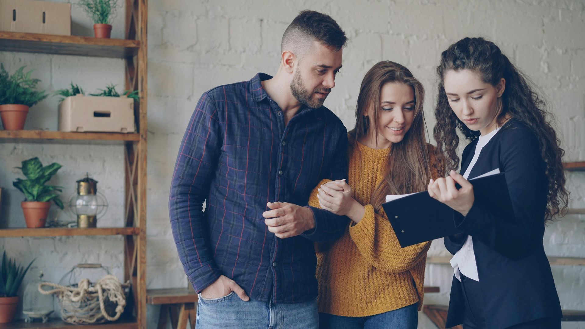 A person in a black blazer reviews a document with a couple, all standing in an indoor setting with shelves.