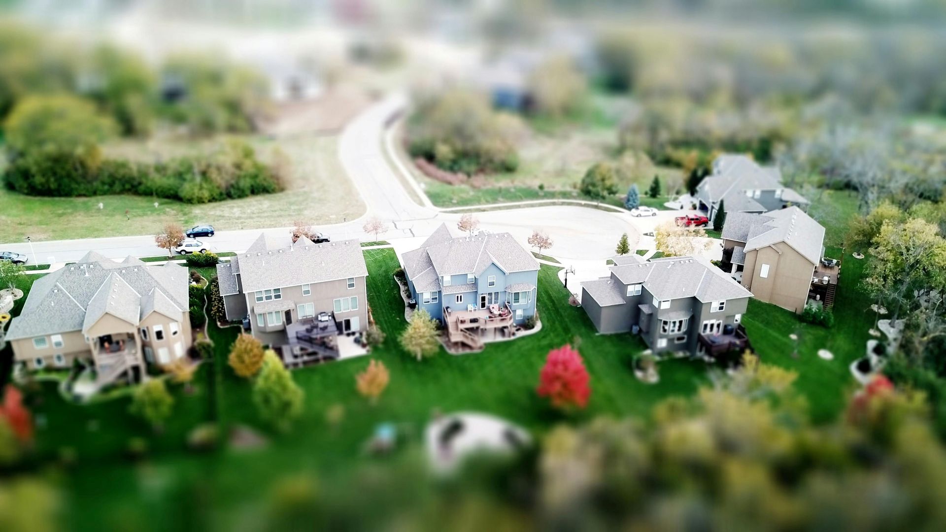 A neighborhood of houses with green lawns and trees, viewed from above with a tilt-shift effect.