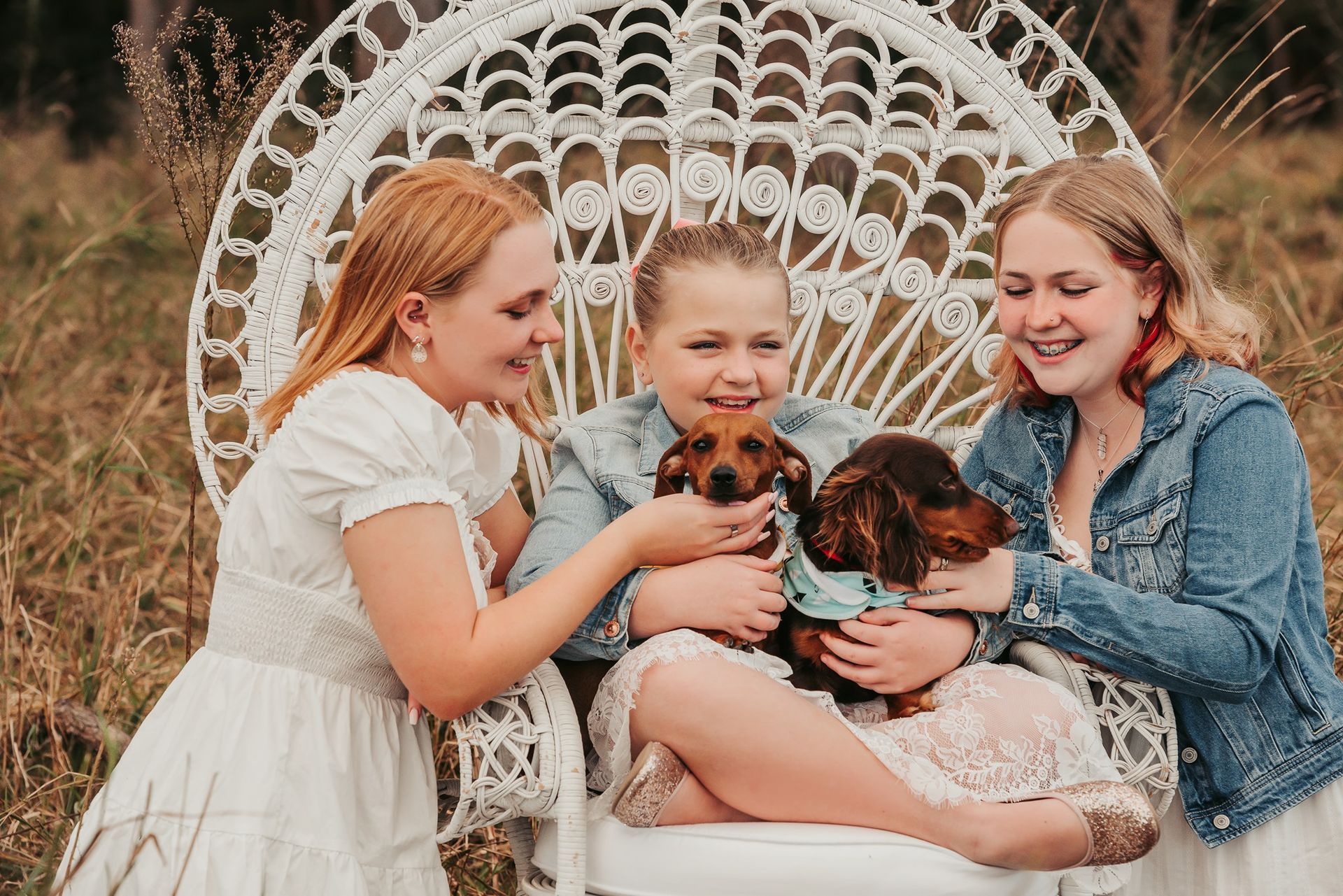 Three people sitting in a white chair hold two dachshund puppies in a field. Smiling, they are dressed in casual attire.
