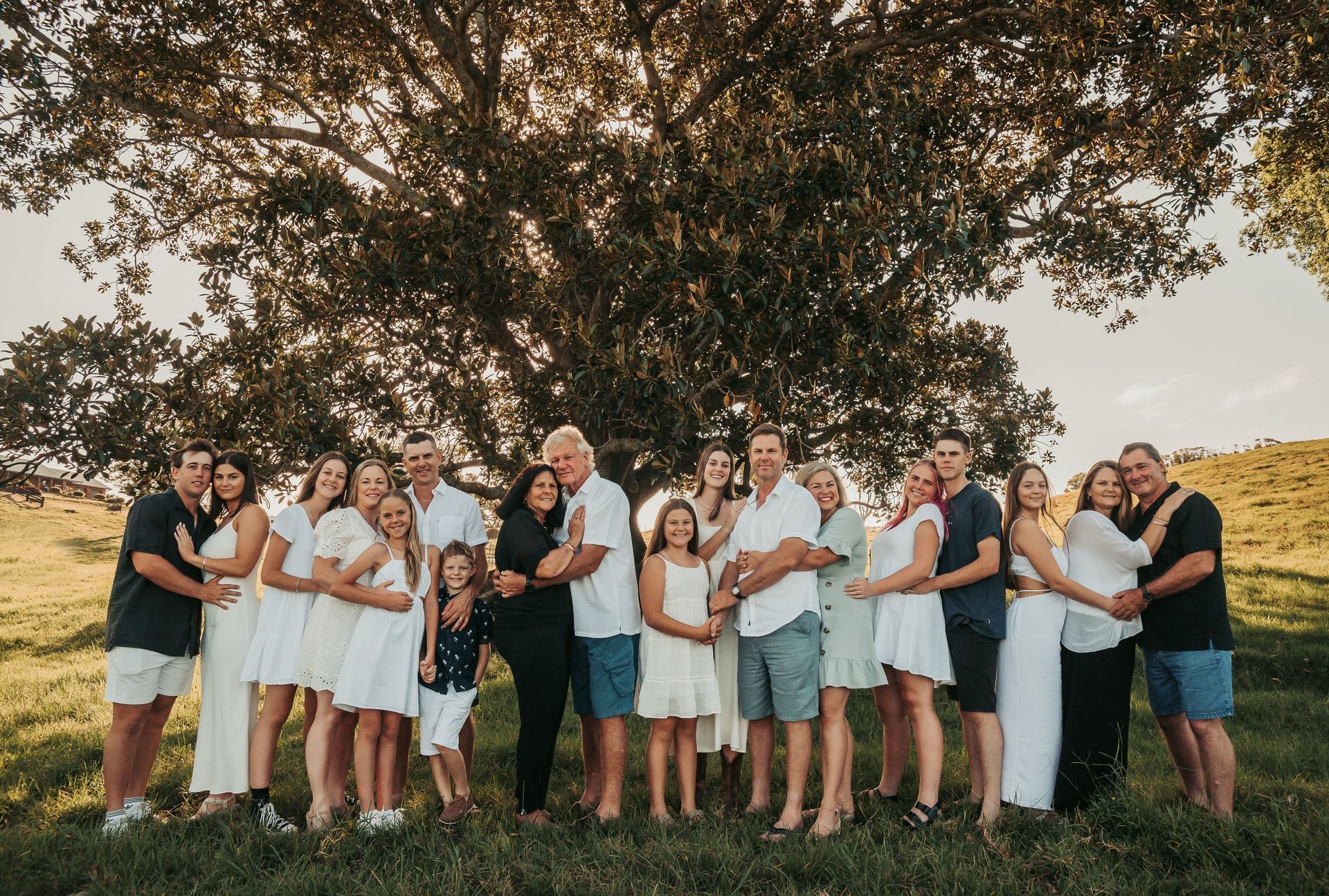 Large family poses for a photo under a tree in a sunny field. People embrace, wearing light colors.