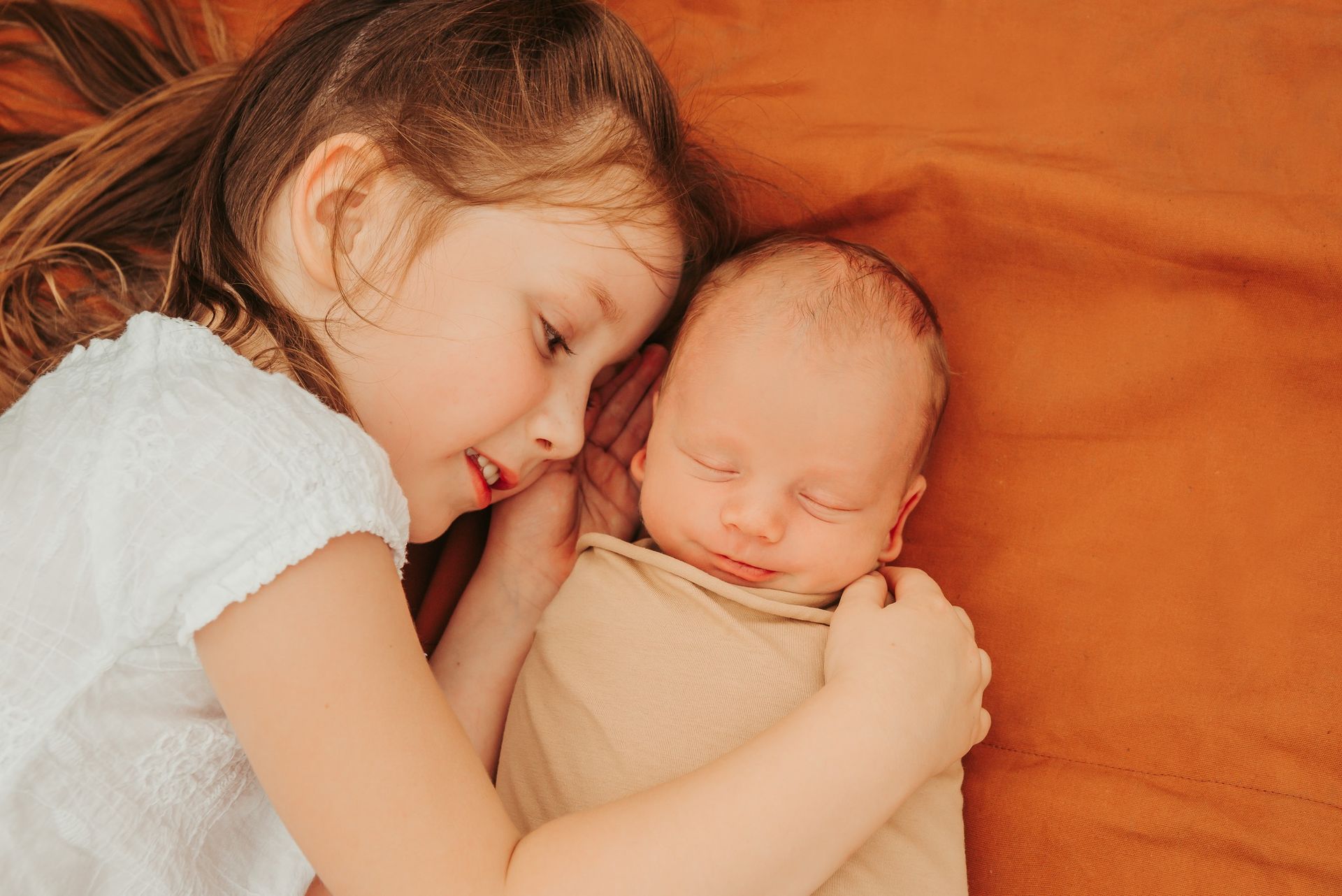 Older Child Smiles, Gently Embracing a Swaddled Infant on an Orange Blanket — Feathers & Lace Photography in Tarzali, QLD