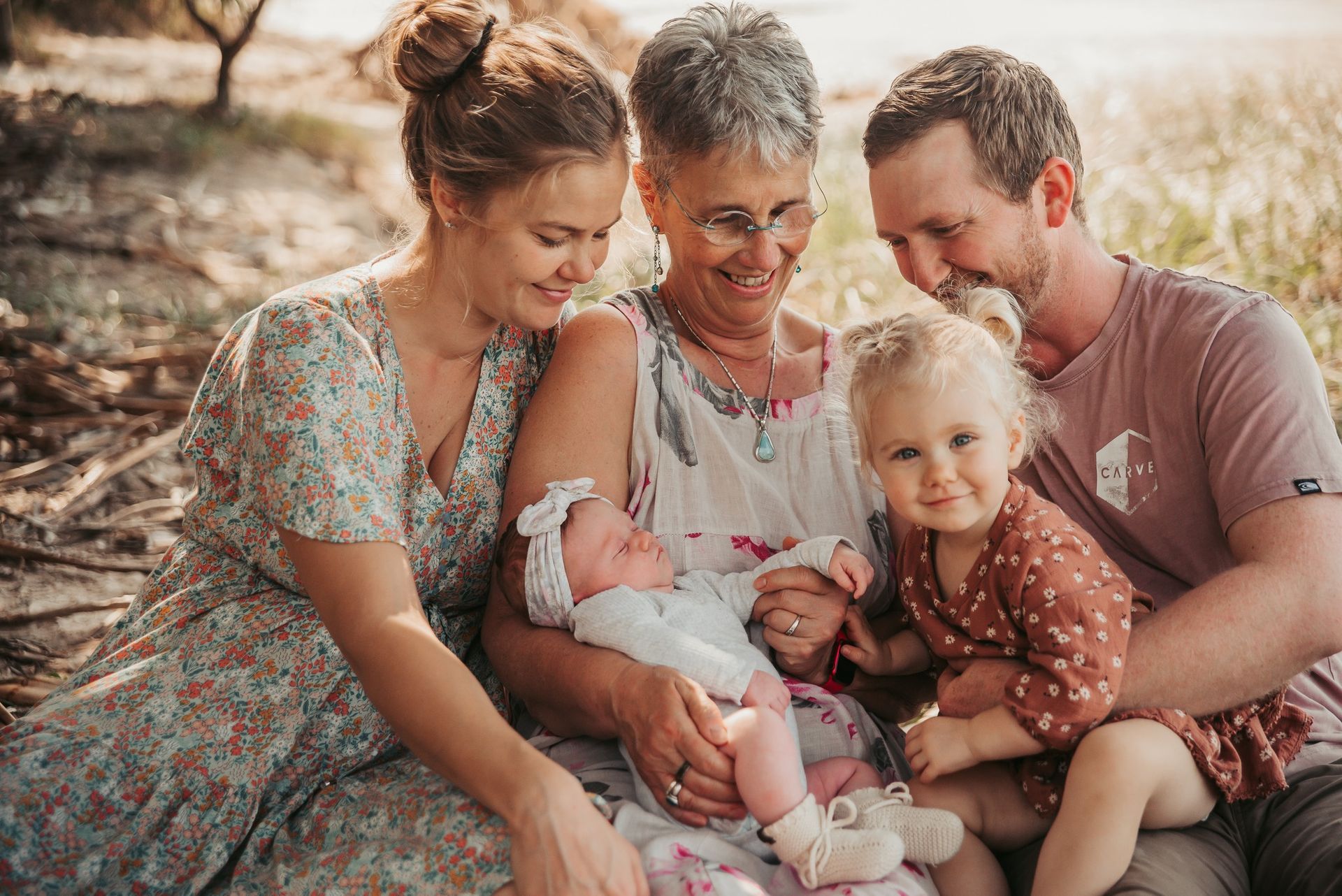 Family of five smiling, gathered outdoors; grandmother holds newborn, other family members look on.