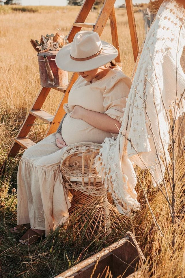 Pregnant Person in Cream Dress and Hat Sits in Wicker Chair in a Field — Feathers & Lace Photography in Tarzali, QLD