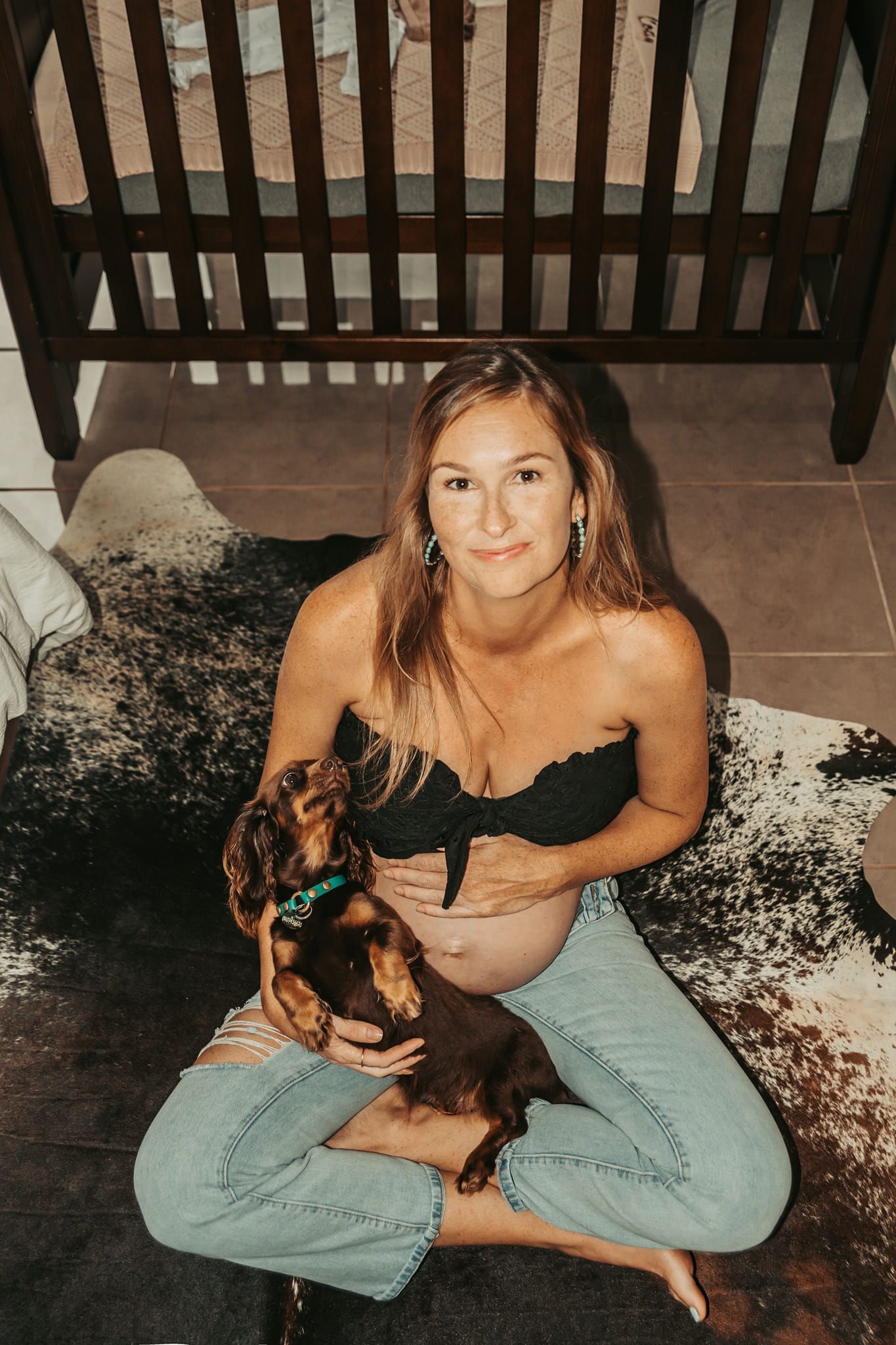 Pregnant Person Seated on a Rug, Holding a Dog, With a Crib in the Background — Feathers & Lace Photography in Tarzali, QLD