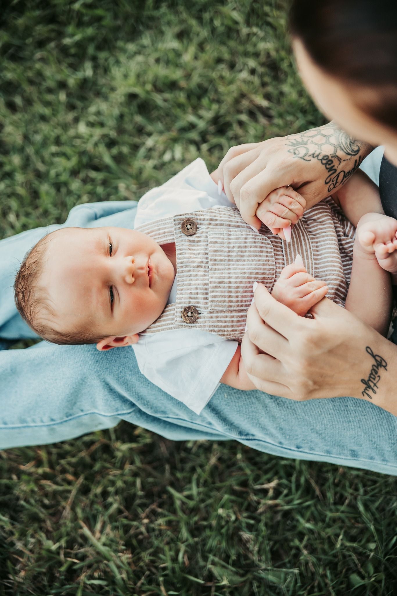 Baby in Striped Outfit, Held on Lap by Person With Tattoos, Resting on Grass — Feathers & Lace Photography in Tarzali, QLD