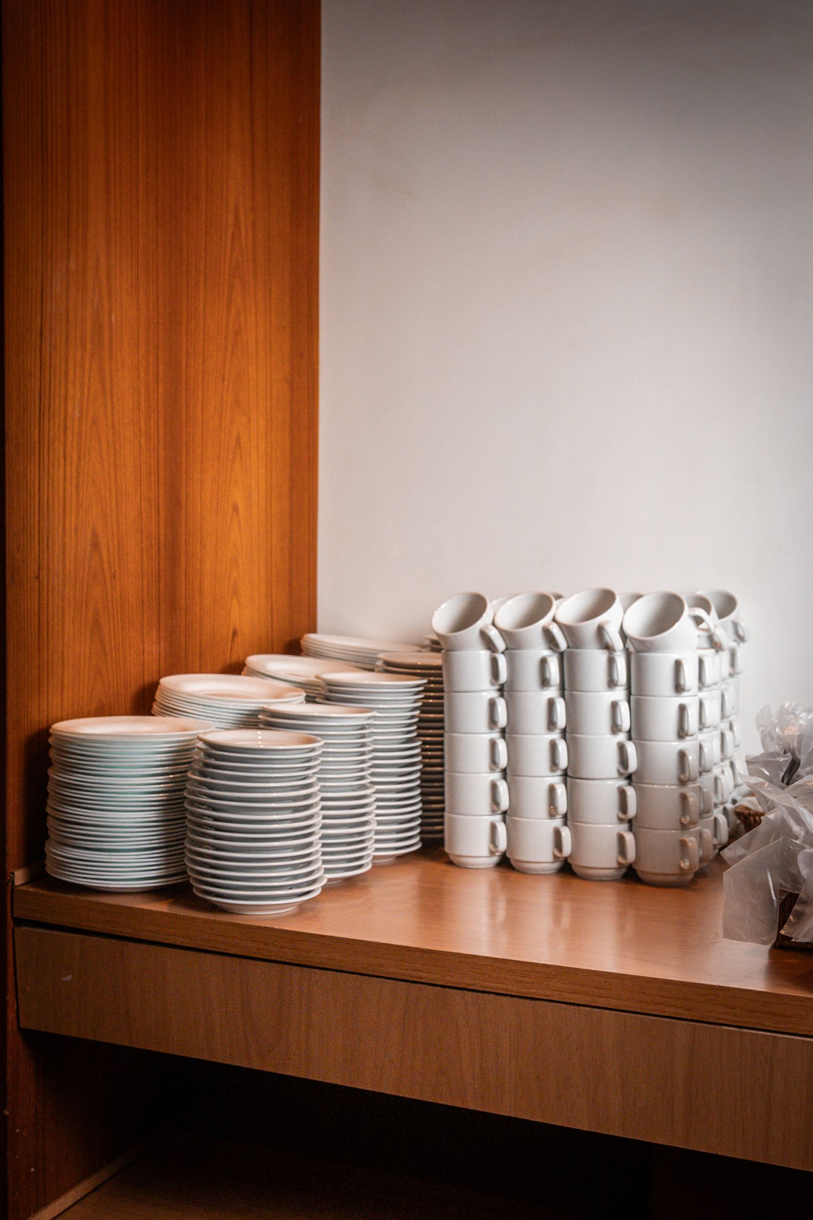 Pilhas de xícaras e pratos brancos sobre uma bancada de madeira, com uma parede de madeira e uma parede branca ao fundo.