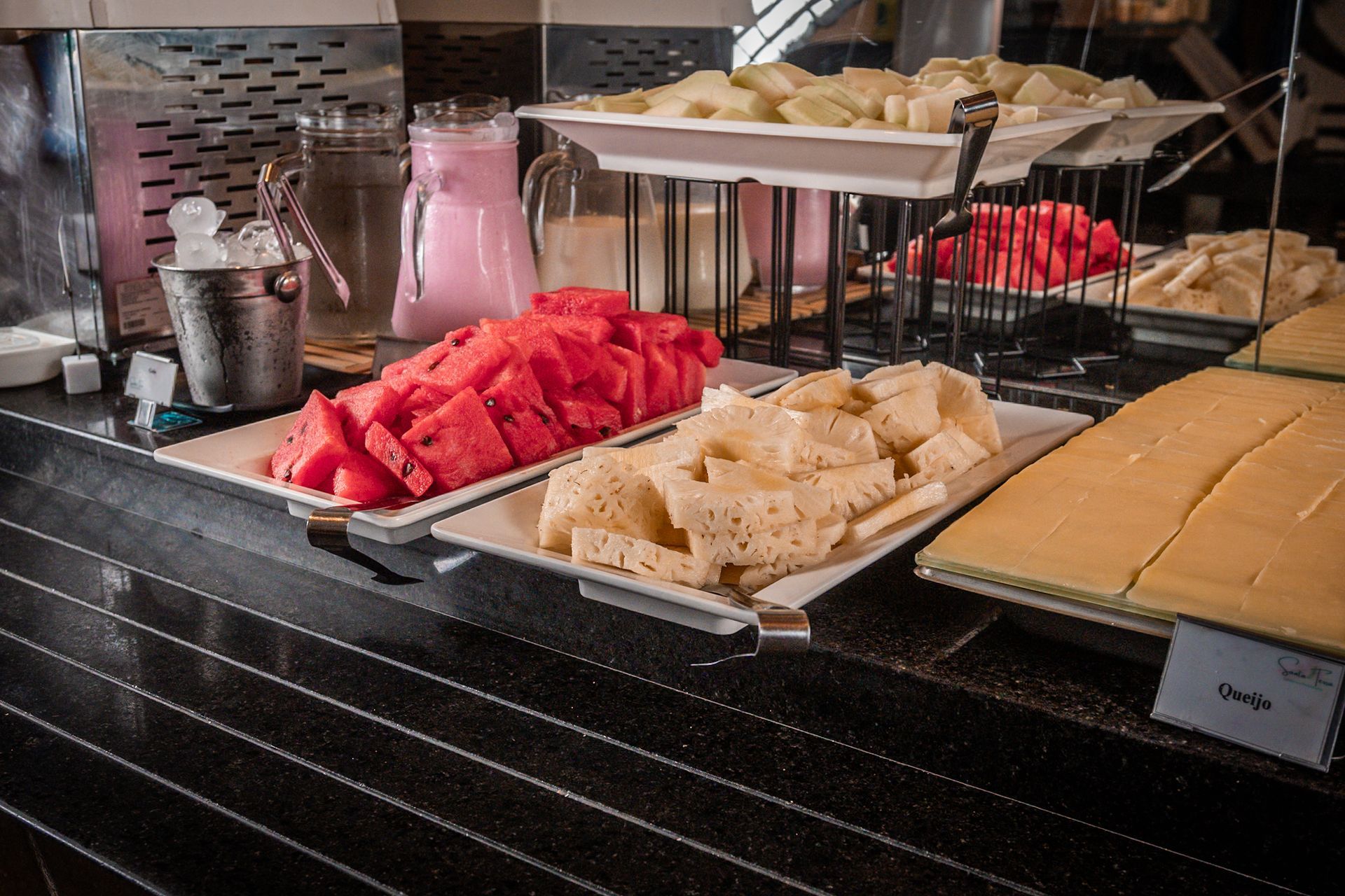 Buffet com melancia, queijo e bebidas rosas e transparentes.