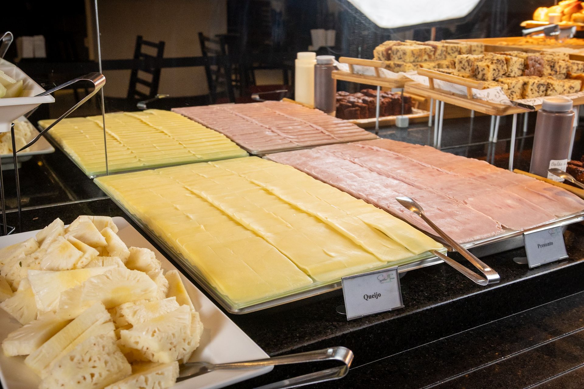 Buffet com fatias de queijo, presunto e pão em exposição.