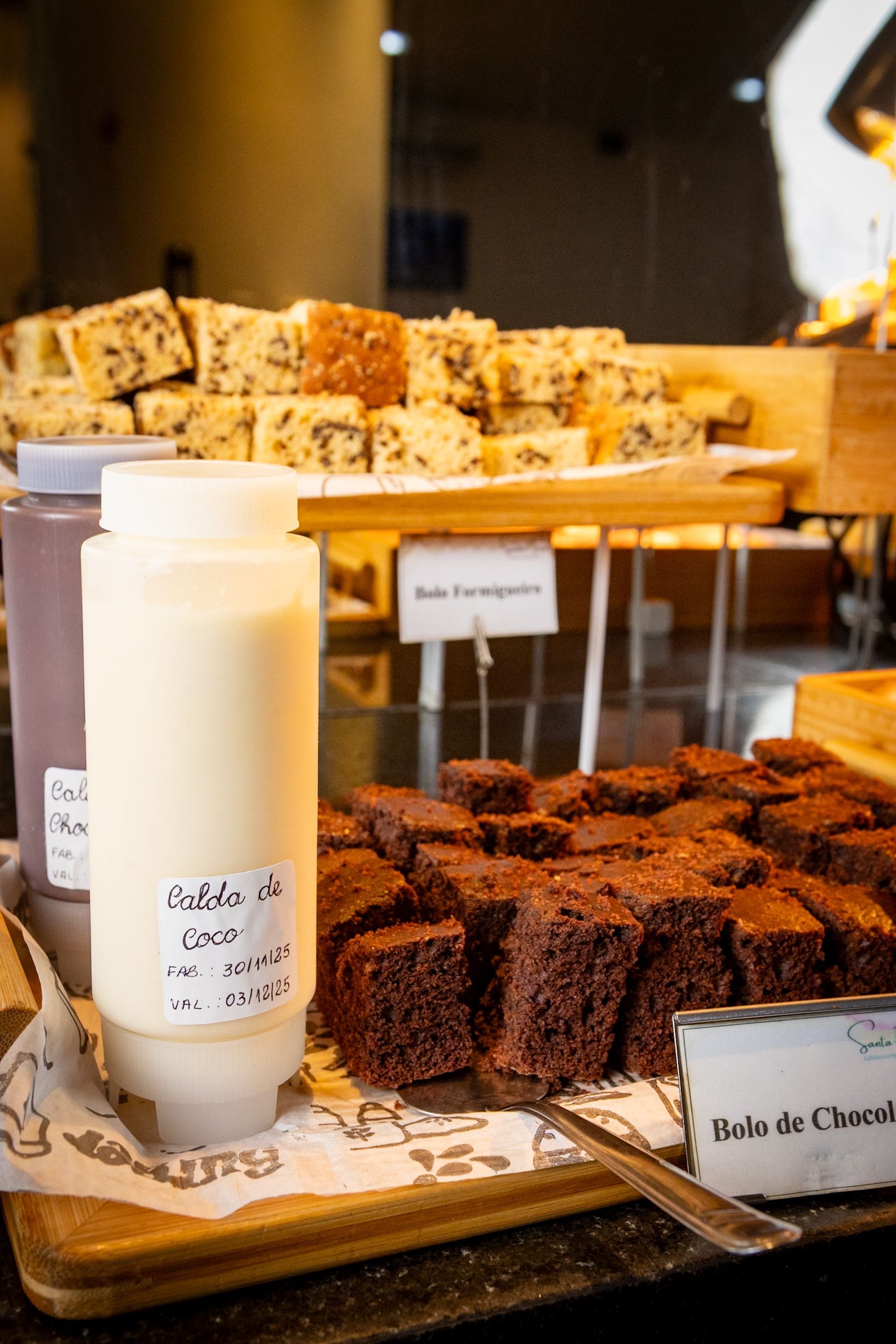 Exposição de sobremesas: brownies, blondies e molhos. Etiquetas indicam os nomes e ingredientes. Prateleiras de madeira.