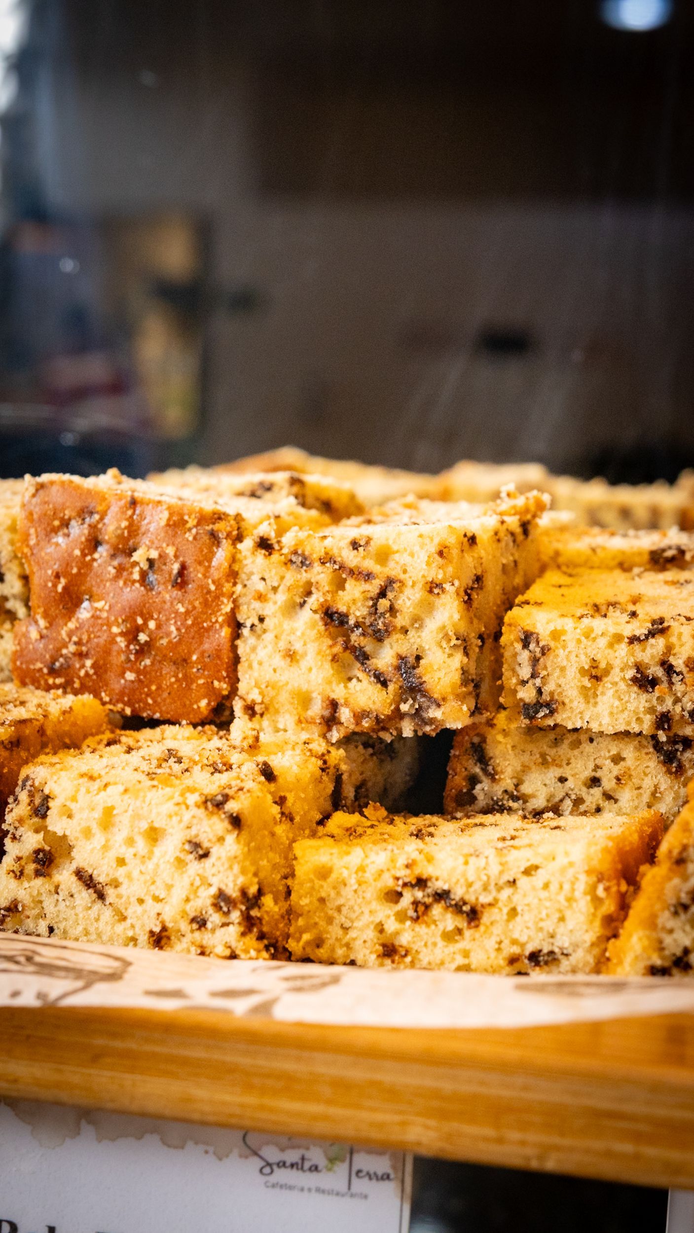 Fatias de bolo com pintas escuras empilhadas sobre uma superfície de madeira, atrás de um vidro.