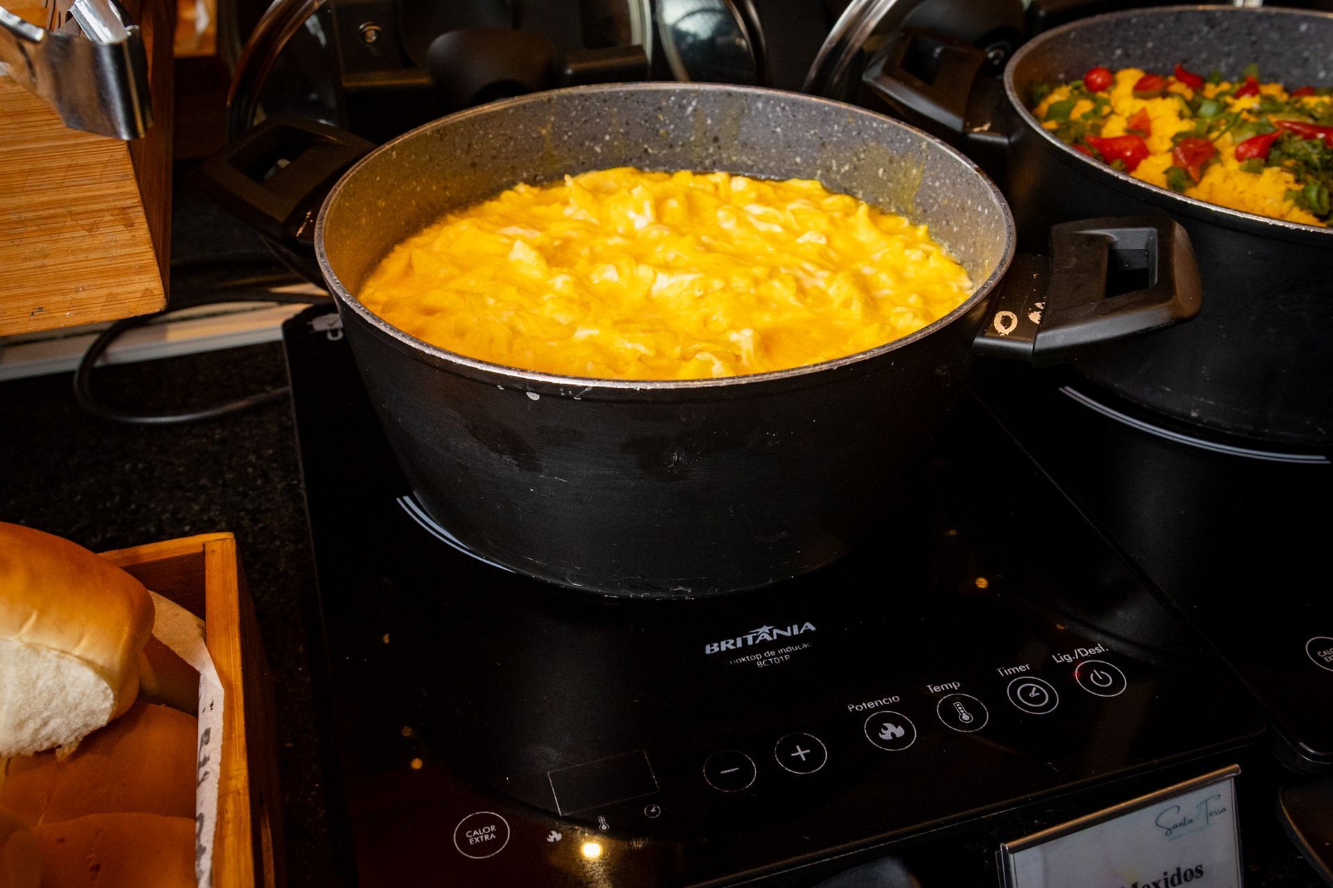 Uma panela com comida cremosa amarela sobre um fogão preto, com uma bandeja de pão à esquerda.
