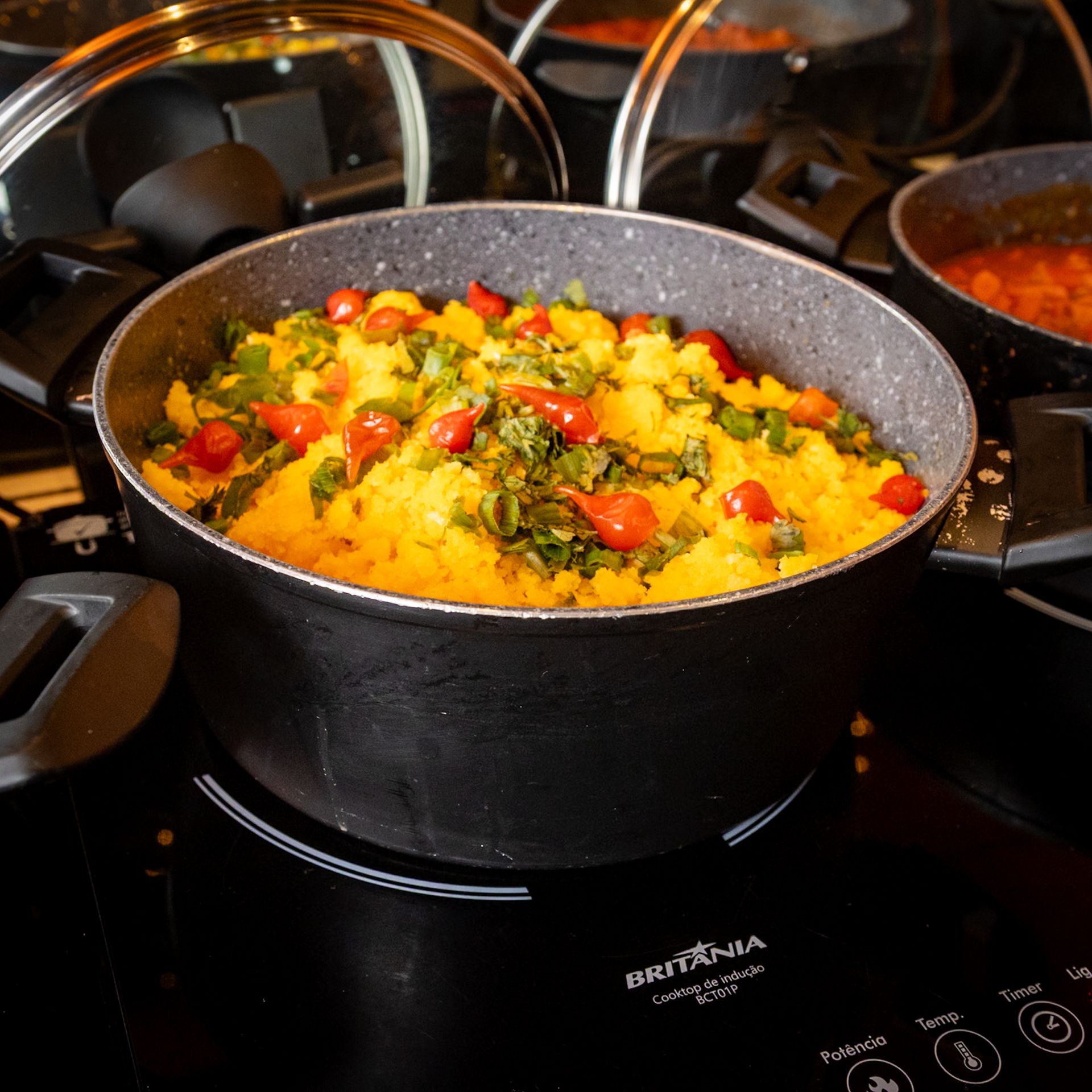 Panela com comida amarela coberta com pimentões vermelhos e ervas verdes sobre um fogão preto.