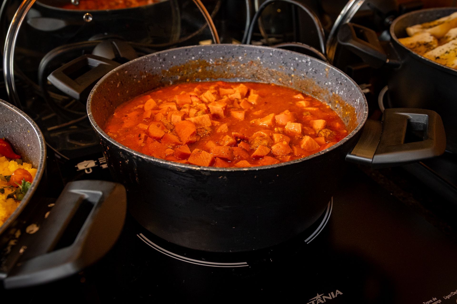 Panela de ensopado com legumes alaranjados em cubos, em molho vermelho, sobre um fogão de chapa preta.