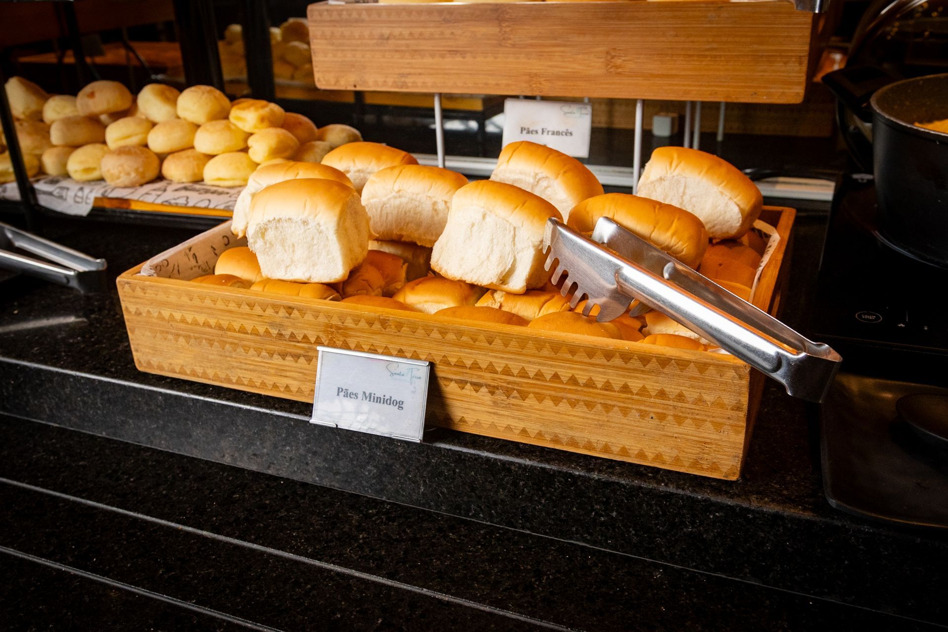 Pãezinhos em uma cesta de madeira com pinças, sobre uma mesa de bufê.
