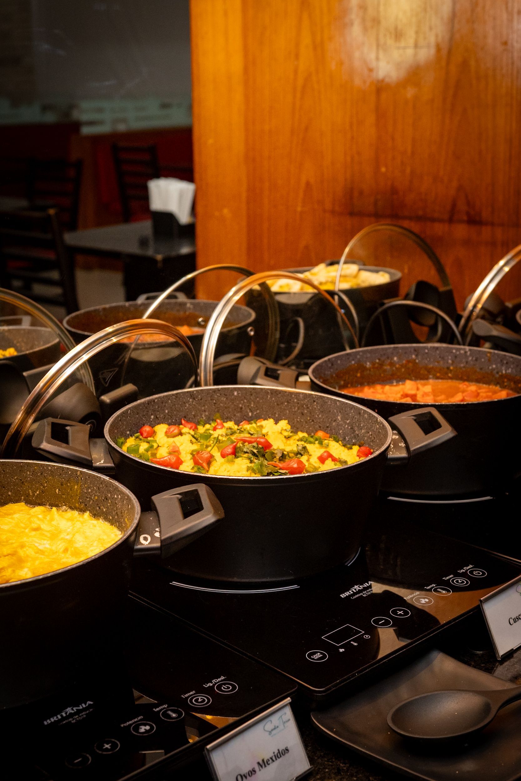 Buffet com diversos pratos em panelas pretas, dispostos em um ambiente de restaurante.