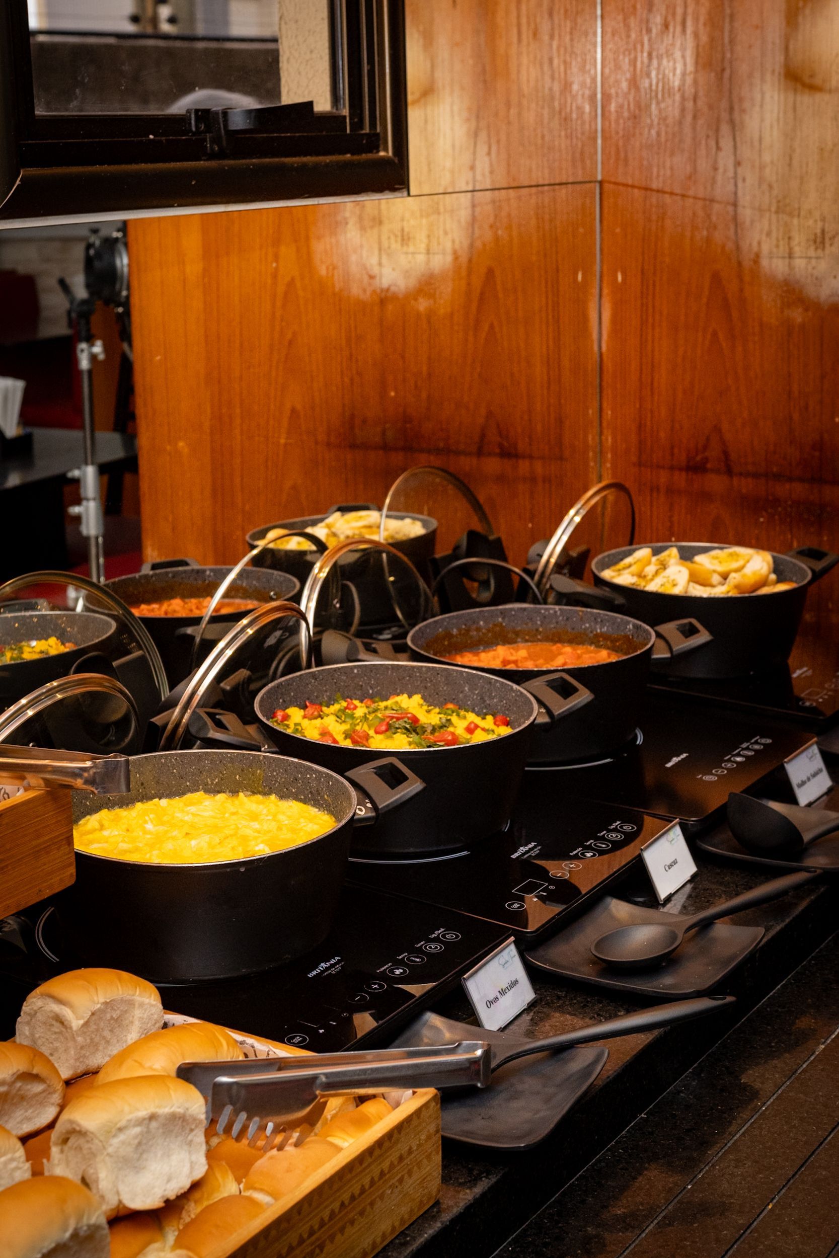 Buffet com várias panelas de ferro fundido contendo diversos itens alimentícios. Pães e utensílios para servir à frente.