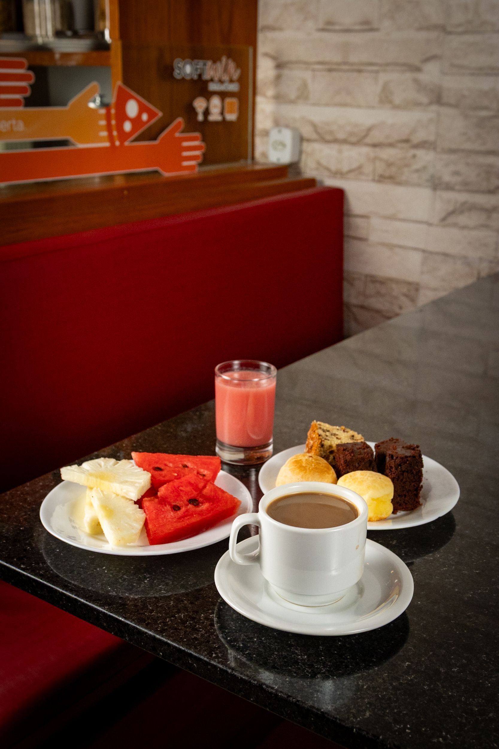 Mesa de café da manhã posta: prato de frutas, café, suco e bolinhos, sobre bancada preta, com mesa e cadeiras vermelhas.