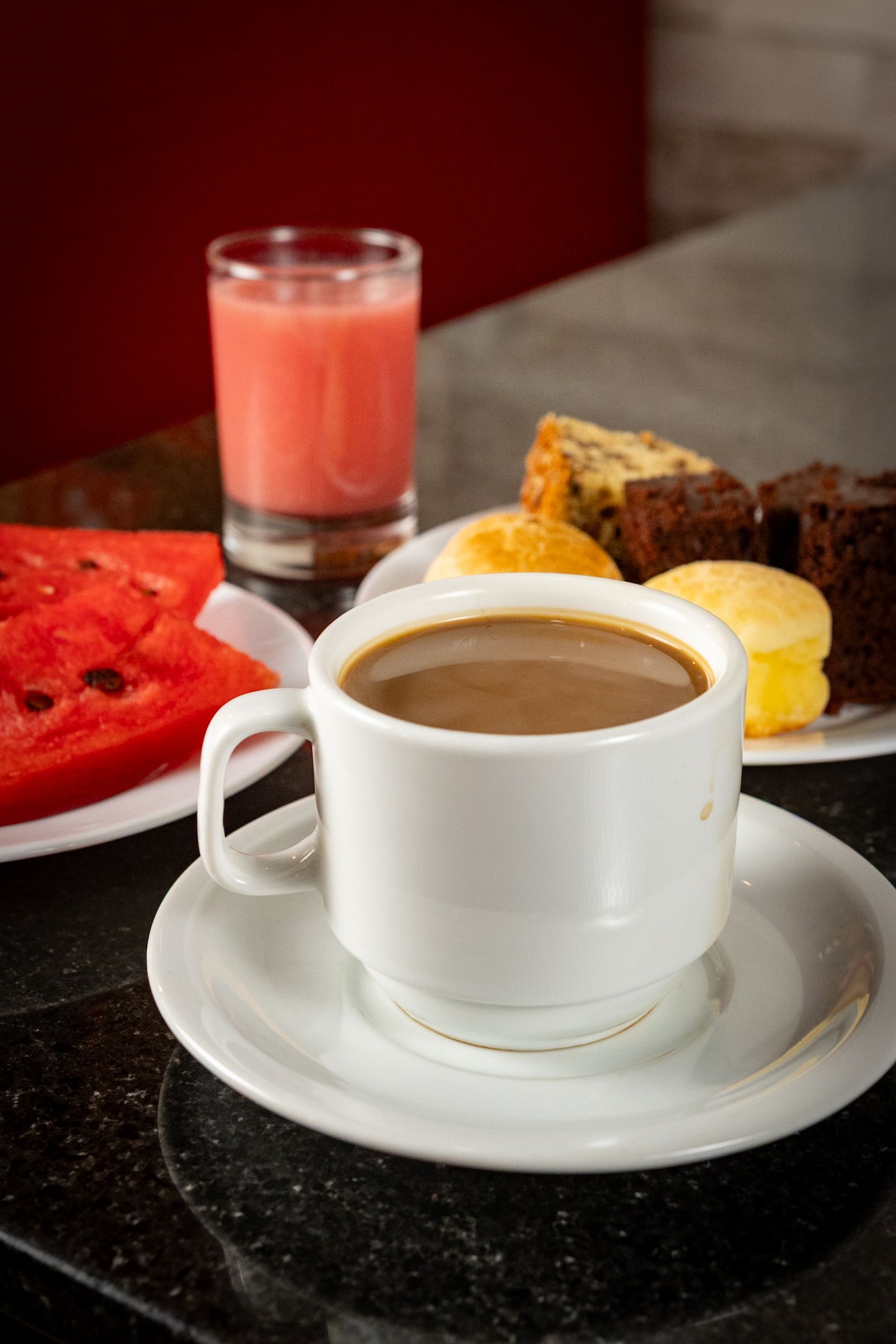 Café em uma xícara branca, melancia, suco e doces sobre a mesa.