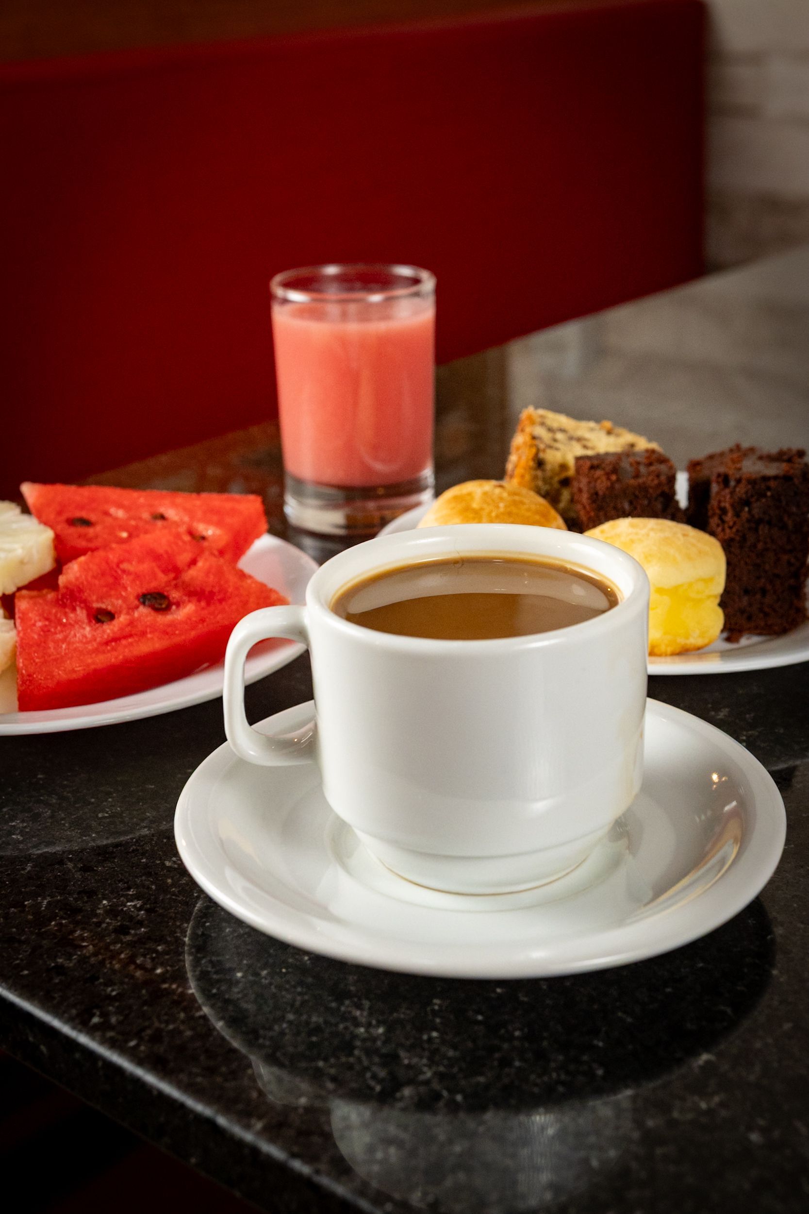 Café em xícara e pires brancos sobre superfície preta, com doces, frutas e suco rosa.