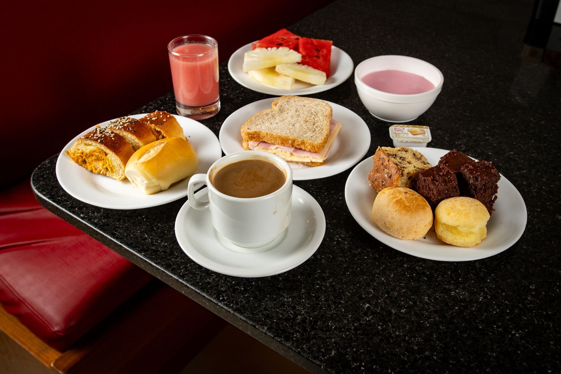 Mesa posta com os seguintes itens de café da manhã: doces, sanduíches, frutas, suco, iogurte e café.