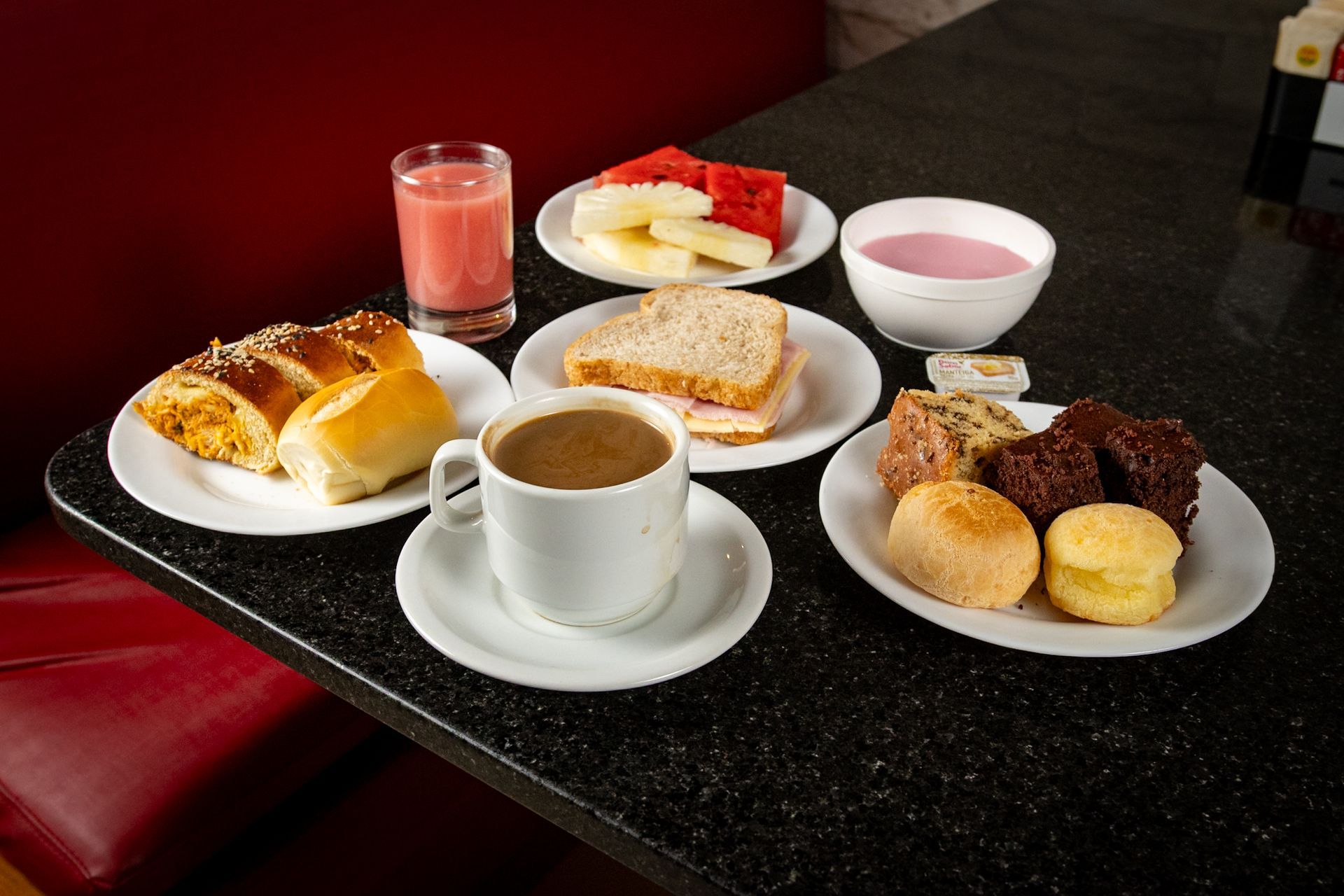 Café da manhã completo com doces, frutas, suco, café e iogurte sobre uma mesa preta com pintas.