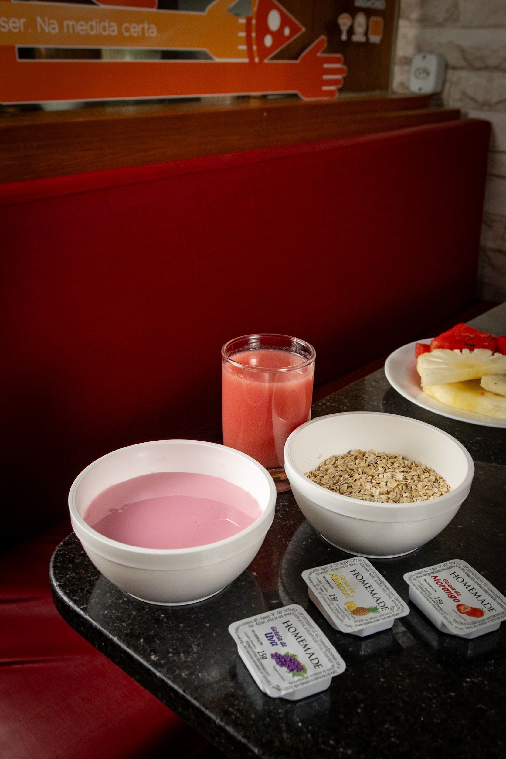 Tigelas de iogurte e cereais, um copo de suco e fatias de frutas sobre uma mesa em uma cabine de lanchonete.