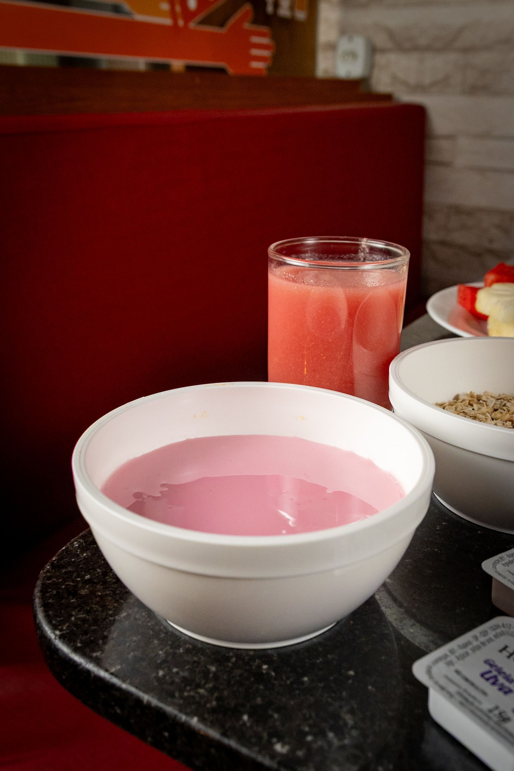 Iogurte rosa em uma tigela branca, copo de suco e outros itens de café da manhã sobre a mesa.