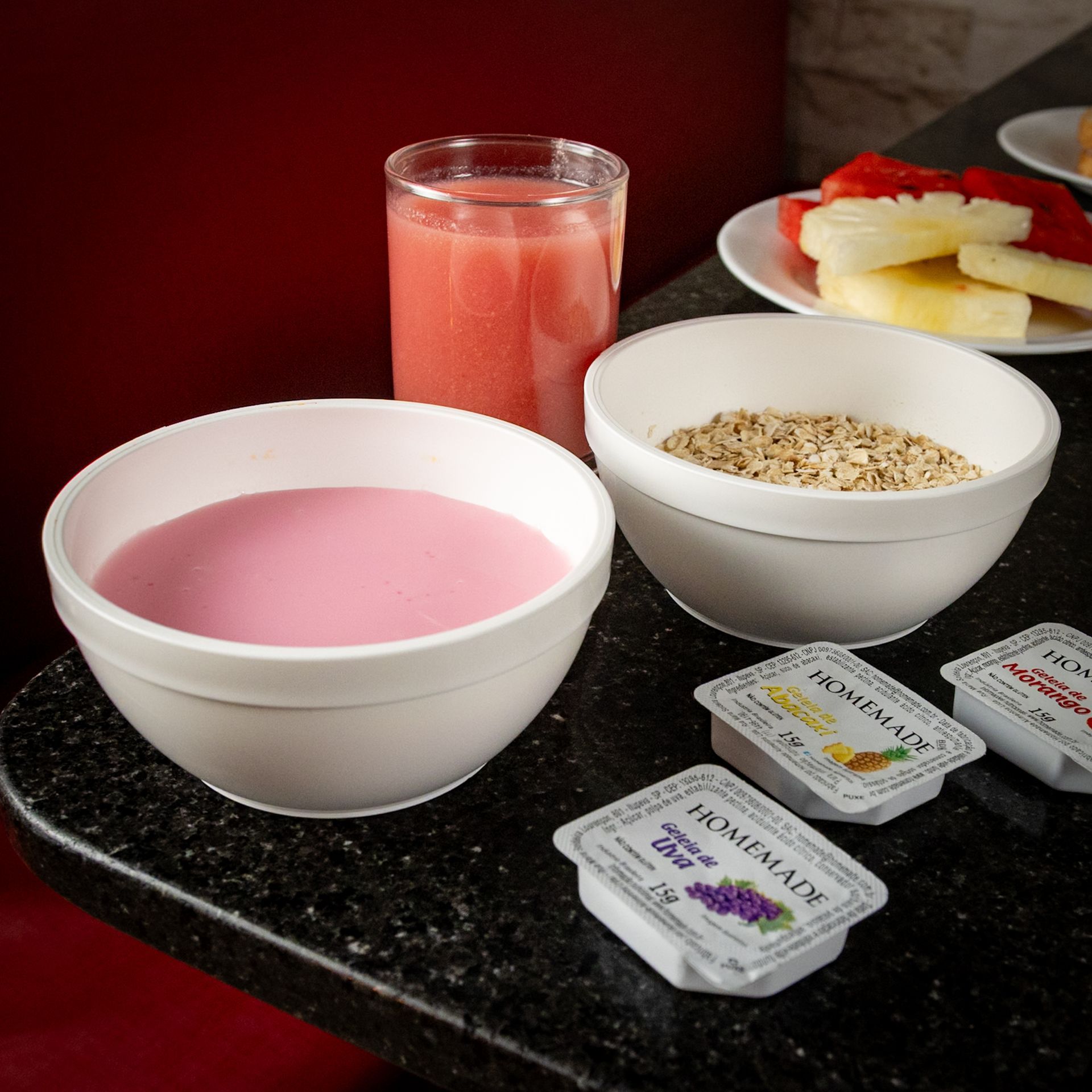 Mesa de café da manhã: iogurte rosa, aveia, frutas, suco e pequenos recipientes sobre uma bancada preta.