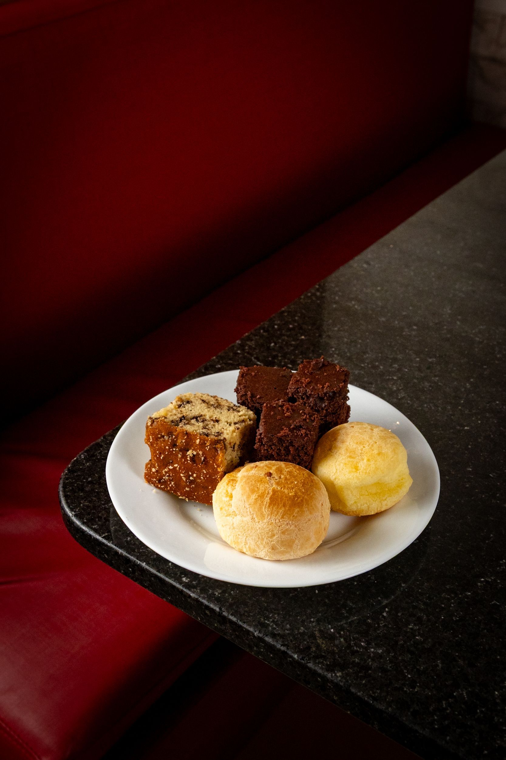 Prato com sobremesas variadas sobre uma mesa escura: brownies, quadradinhos e pãezinhos.