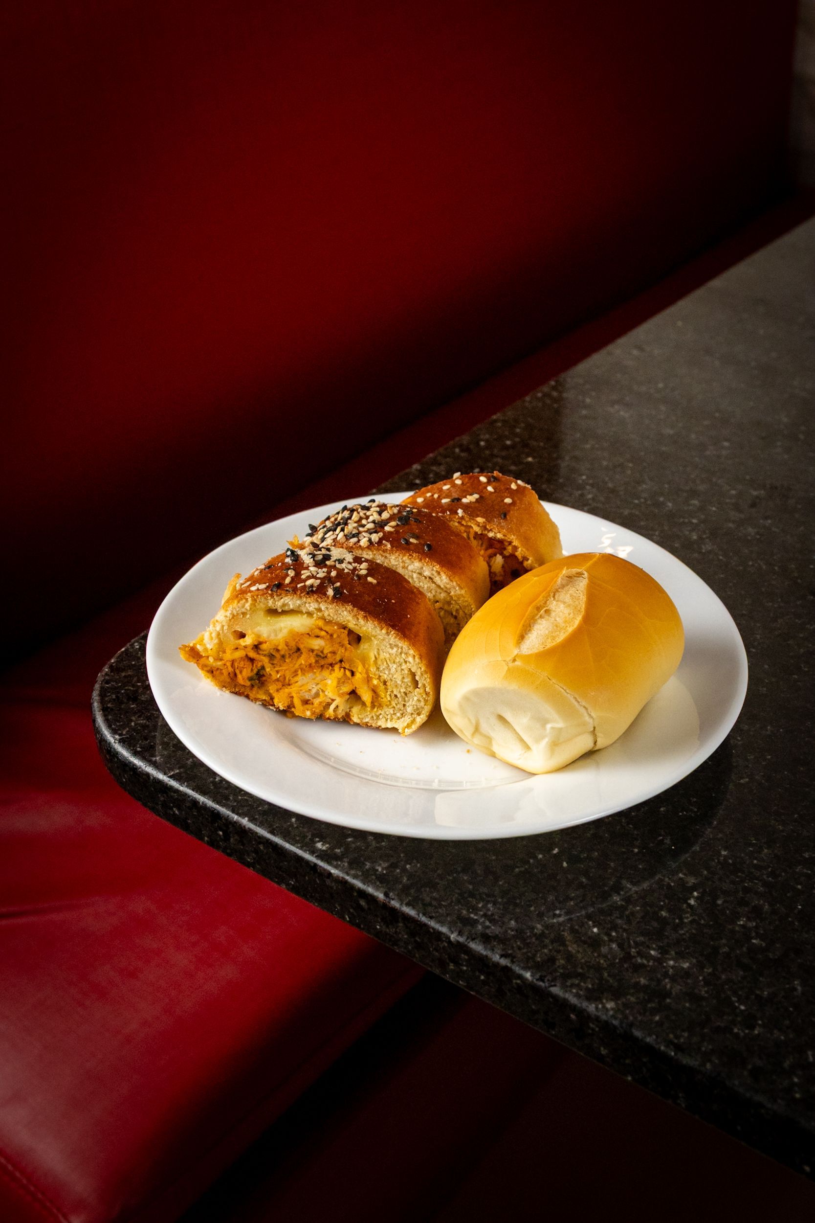 Prato com uma massa parcialmente cortada, recheada com o que parece ser frango, e um pequeno pãozinho sobre uma bancada preta, em um banco vermelho.