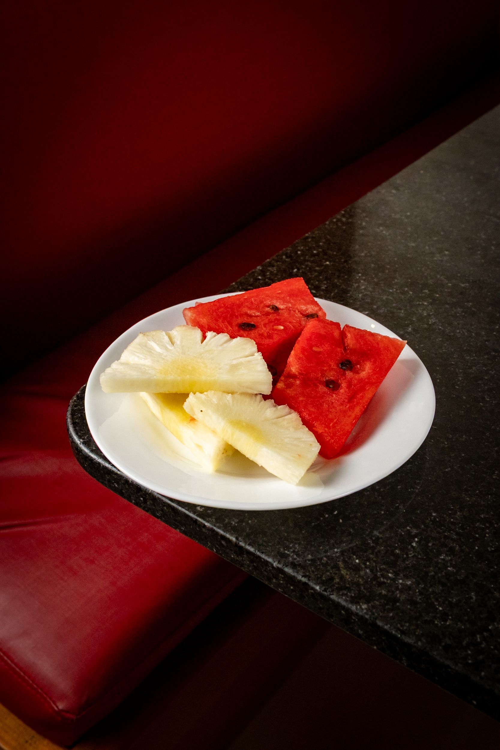 Prato com fatias de melancia e abacaxi sobre uma bancada preta perto de uma mesa vermelha.