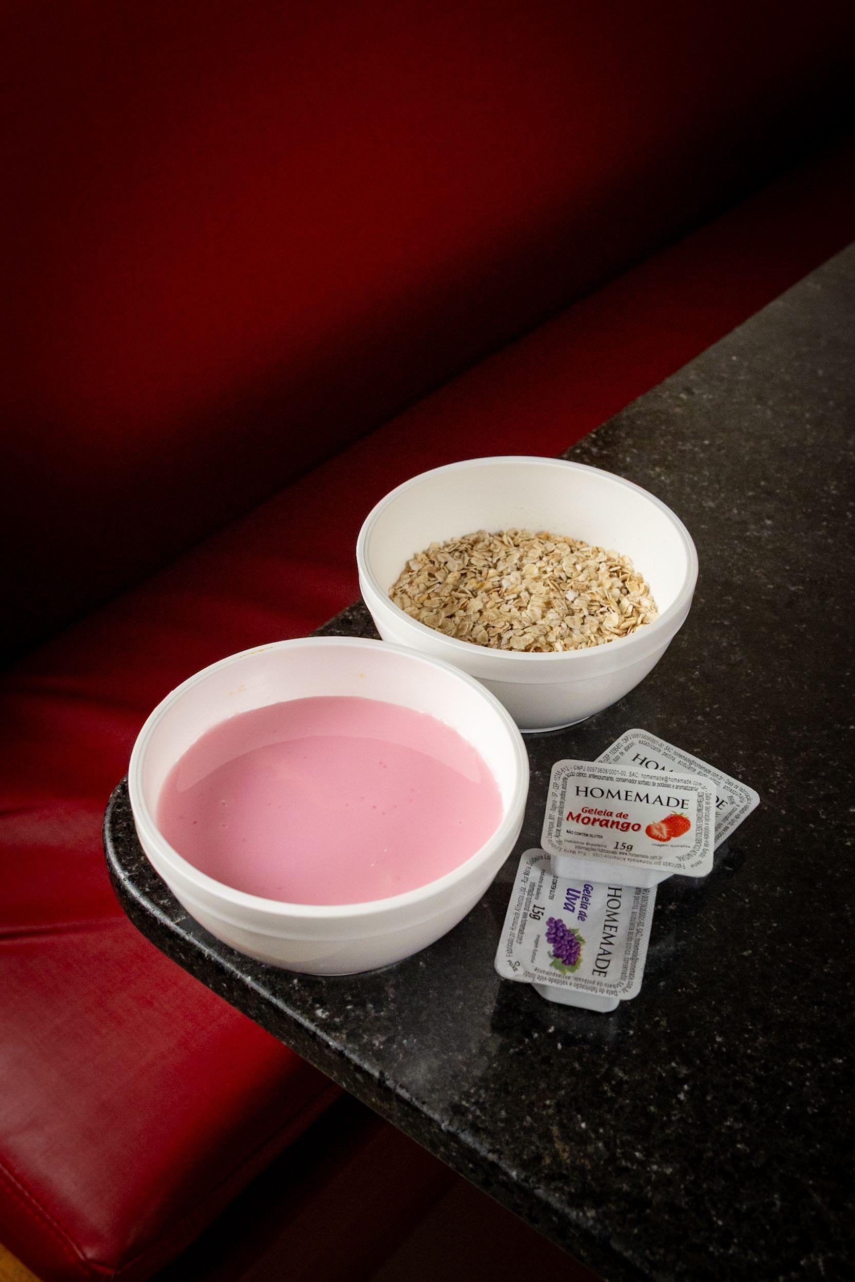 Duas tigelas brancas: uma com líquido rosa, a outra com grãos marrons, e pequenos pacotes sobre um balcão preto perto de uma cabine vermelha.