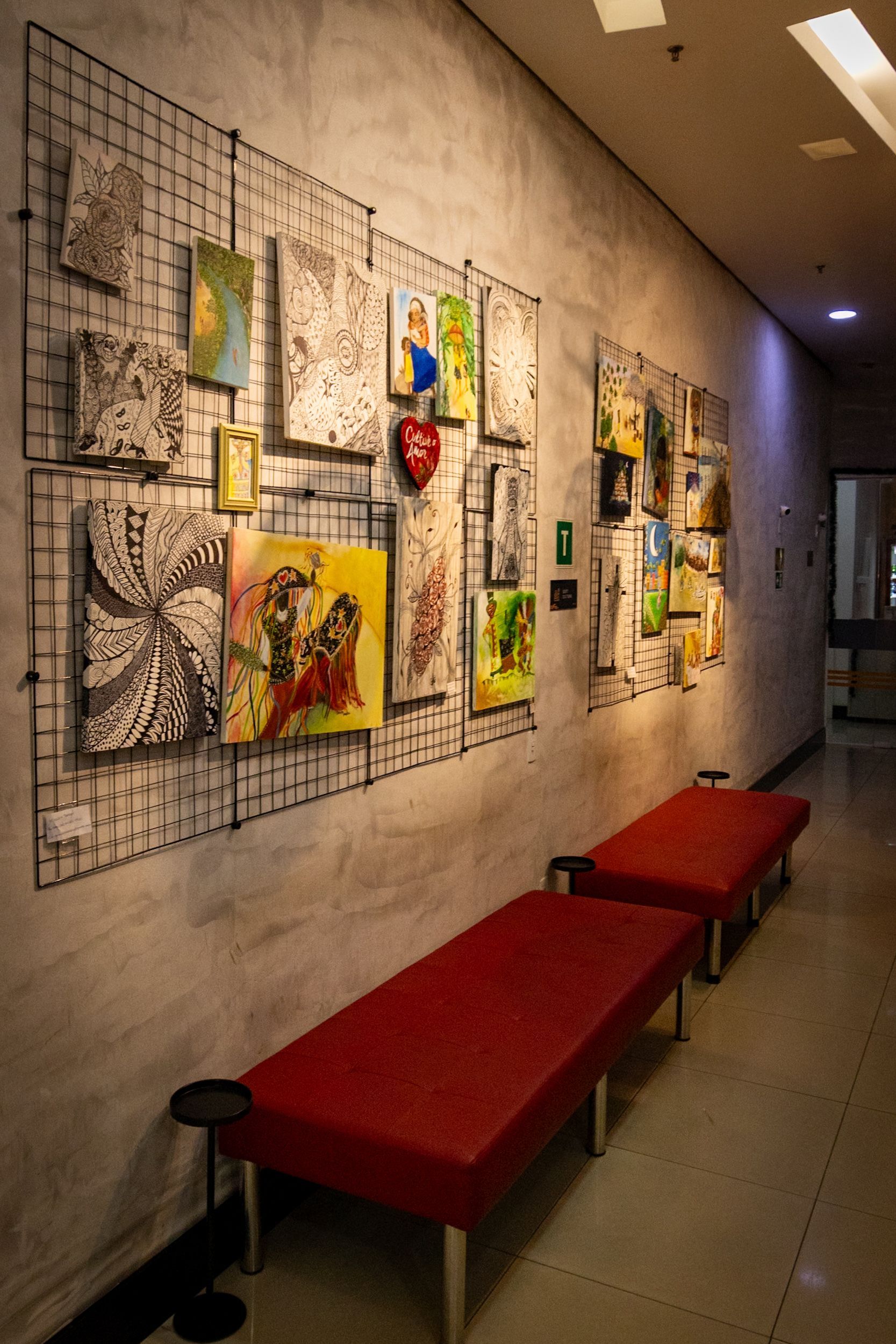 Obras de arte expostas em uma grade de arame contra uma parede de concreto, bancos vermelhos no corredor de uma galeria.