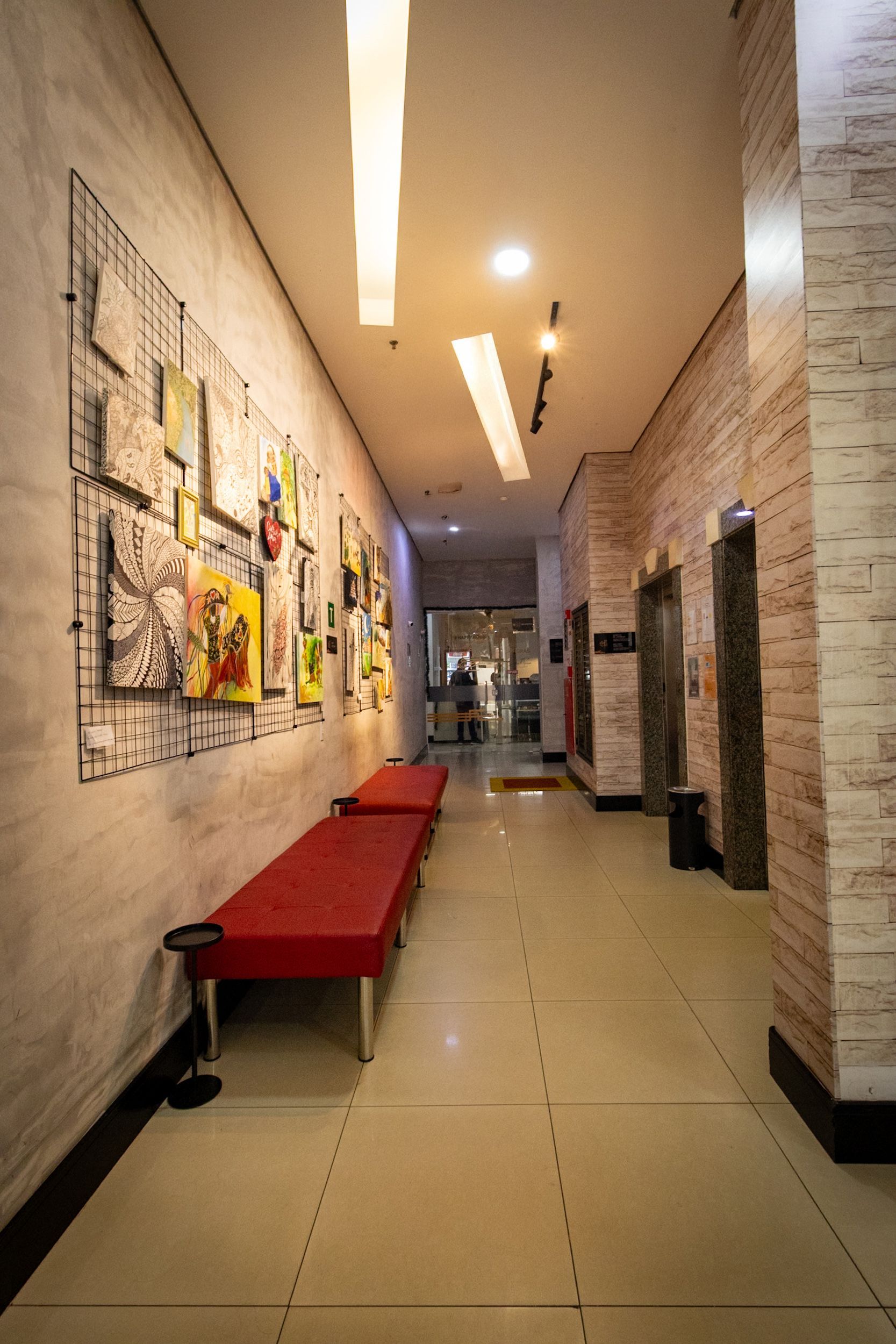 Corredor com banco vermelho, obras de arte na parede e elevadores. Tijolo e azulejo bege com iluminação no teto.