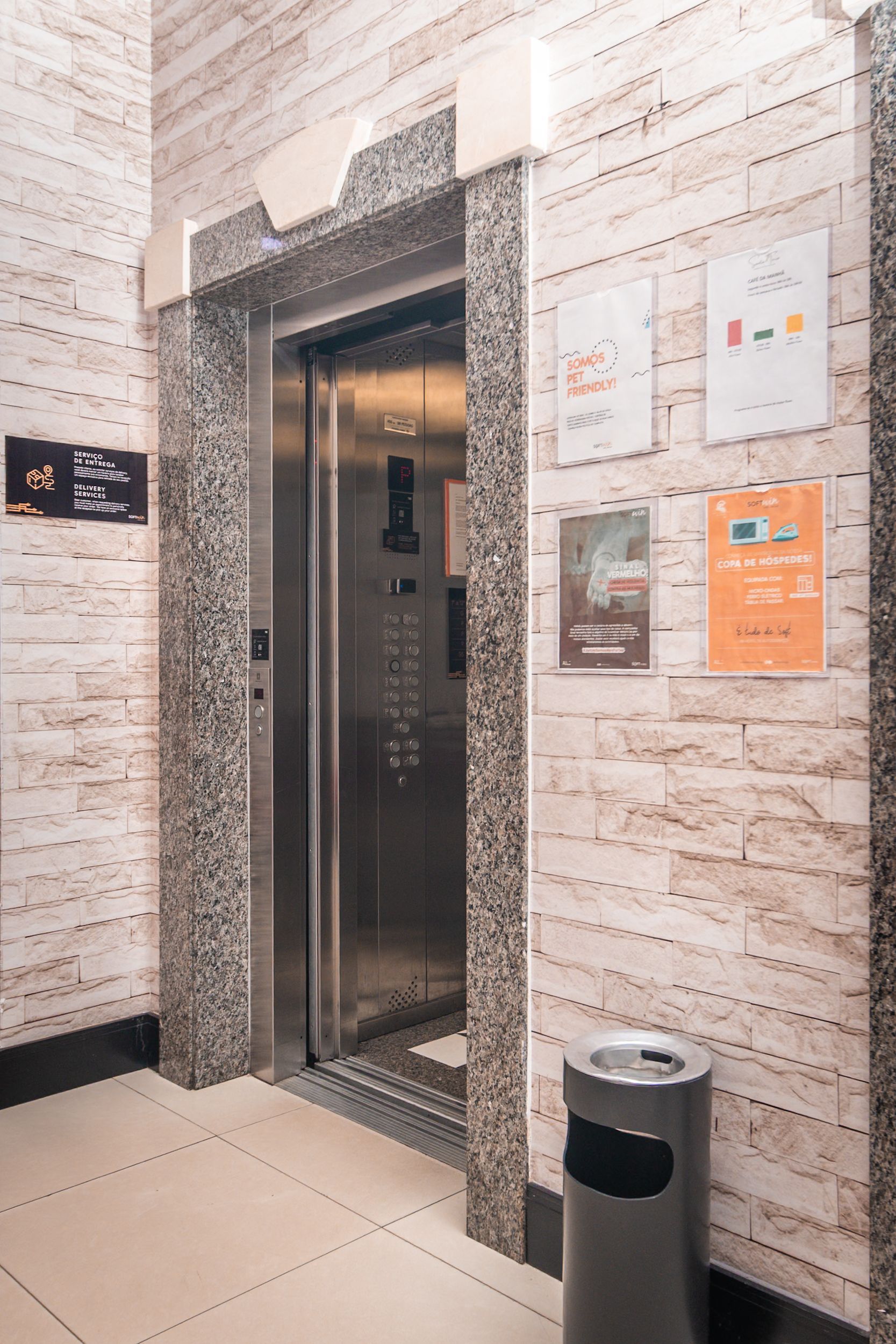 Elevador com portas fechadas de aço inoxidável, emolduradas em mármore. Parede de tijolos bege. Lixeira. Placas.