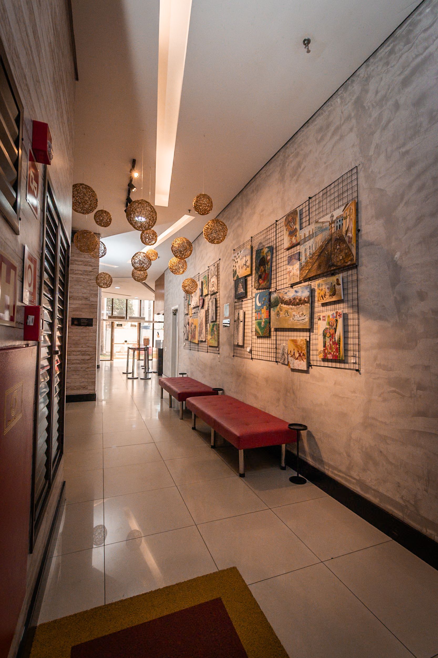 Corredor com obras de arte, bancos vermelhos, azulejos brancos e luminárias esféricas.