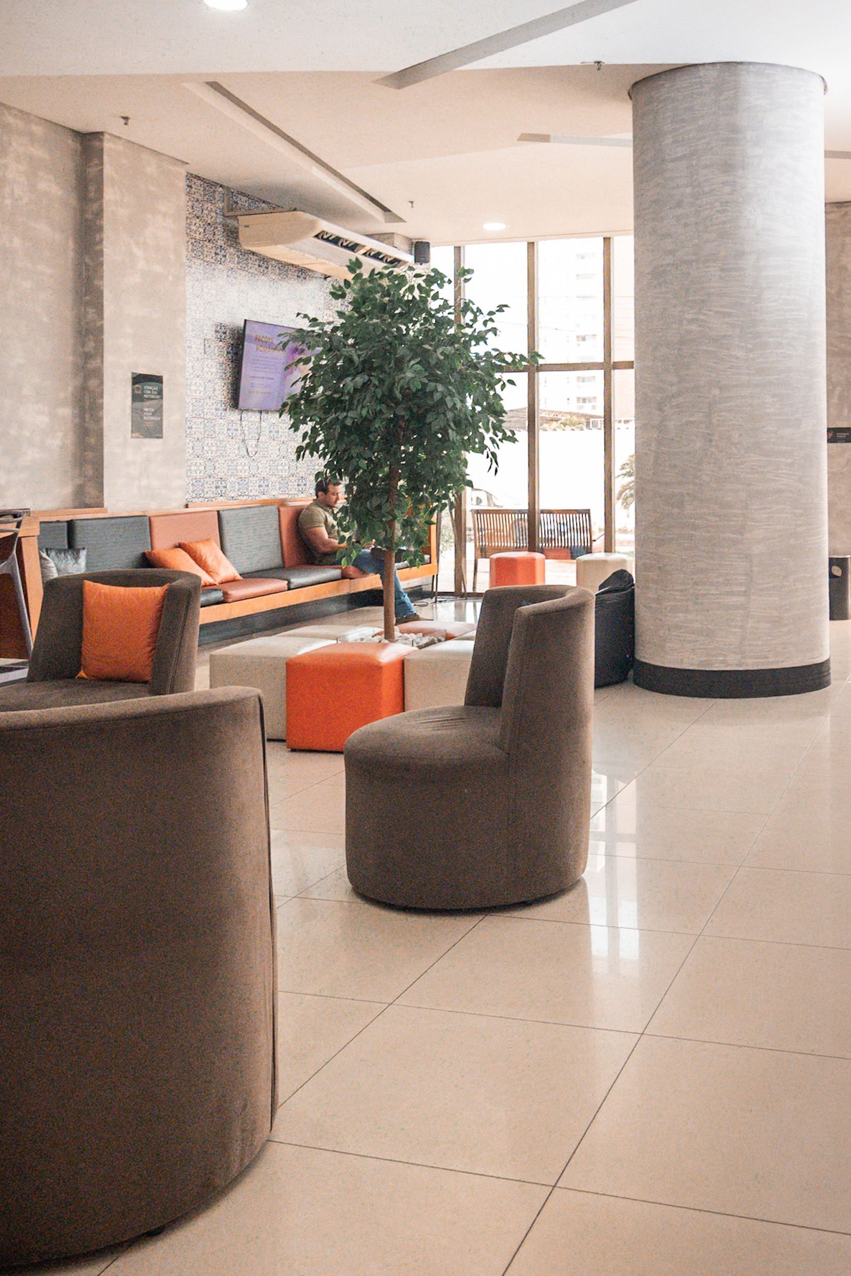 Hall de entrada com assentos, uma planta e uma grande coluna. Detalhes em marrom e laranja com janelas amplas.