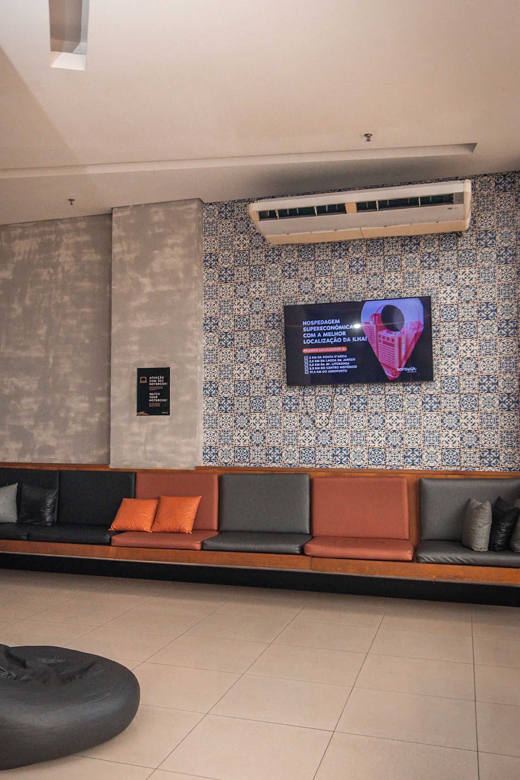 Área de estar com assentos embutidos, parede estampada, TV e pufe. Tons de laranja, cinza e azul.