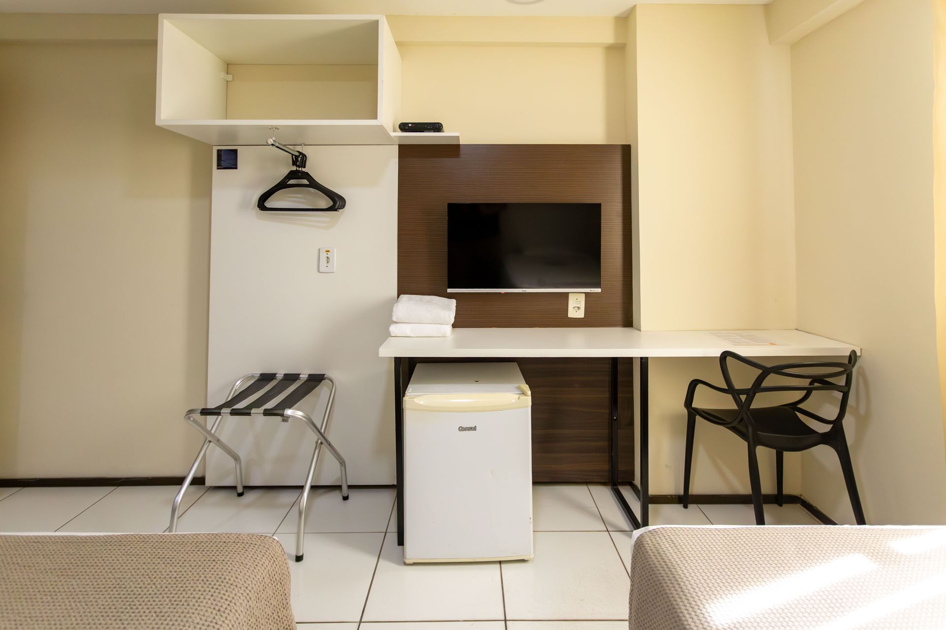 Interior de quarto de hotel com escrivaninha, TV, armário, cadeira, frigobar e suporte para bagagem.