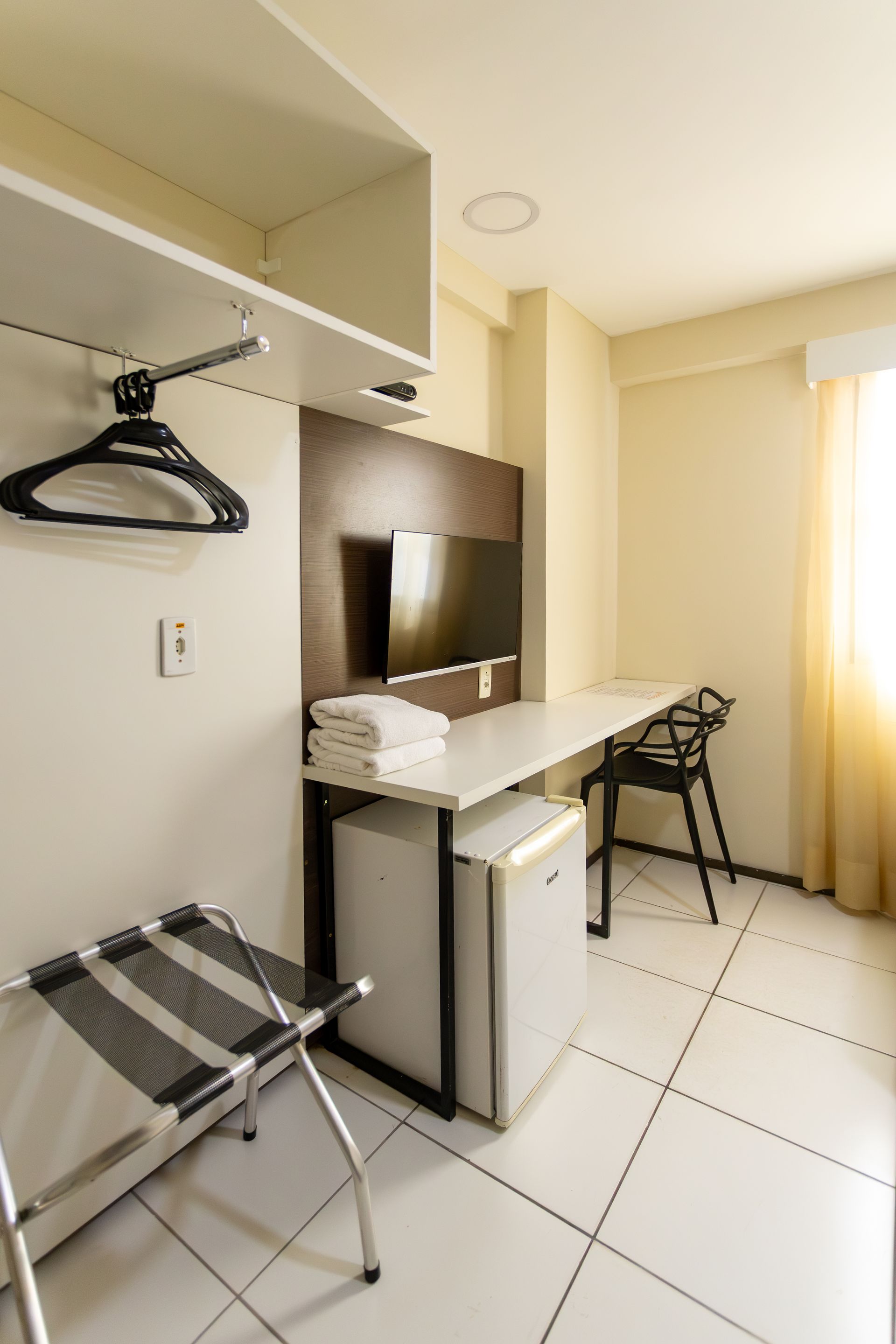 Quarto de hotel com escrivaninha, TV, frigobar e suporte para malas. Azulejos brancos e paredes bege.