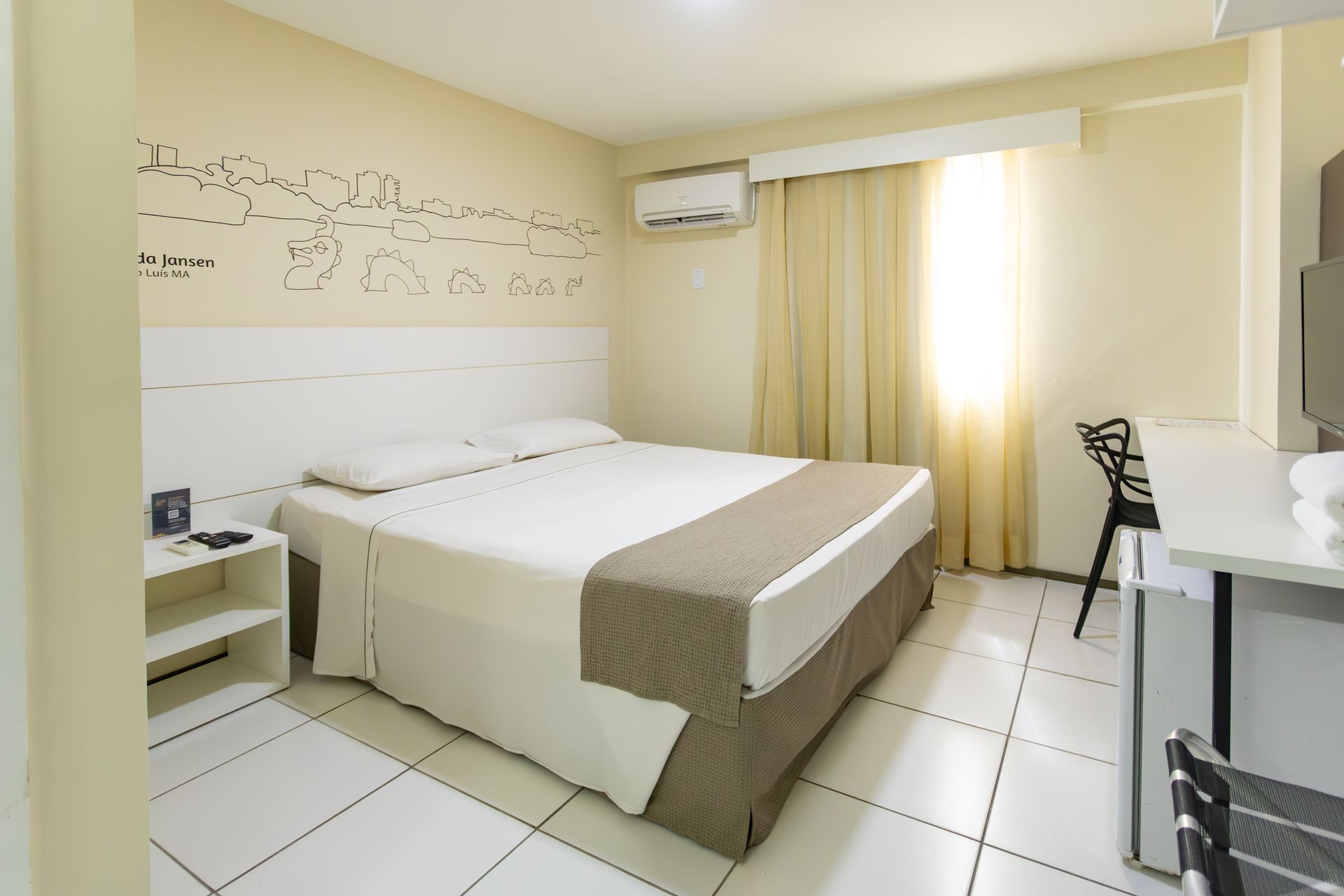 Quarto de hotel com cama de casal, escrivaninha e janela com entrada de luz pelas cortinas.