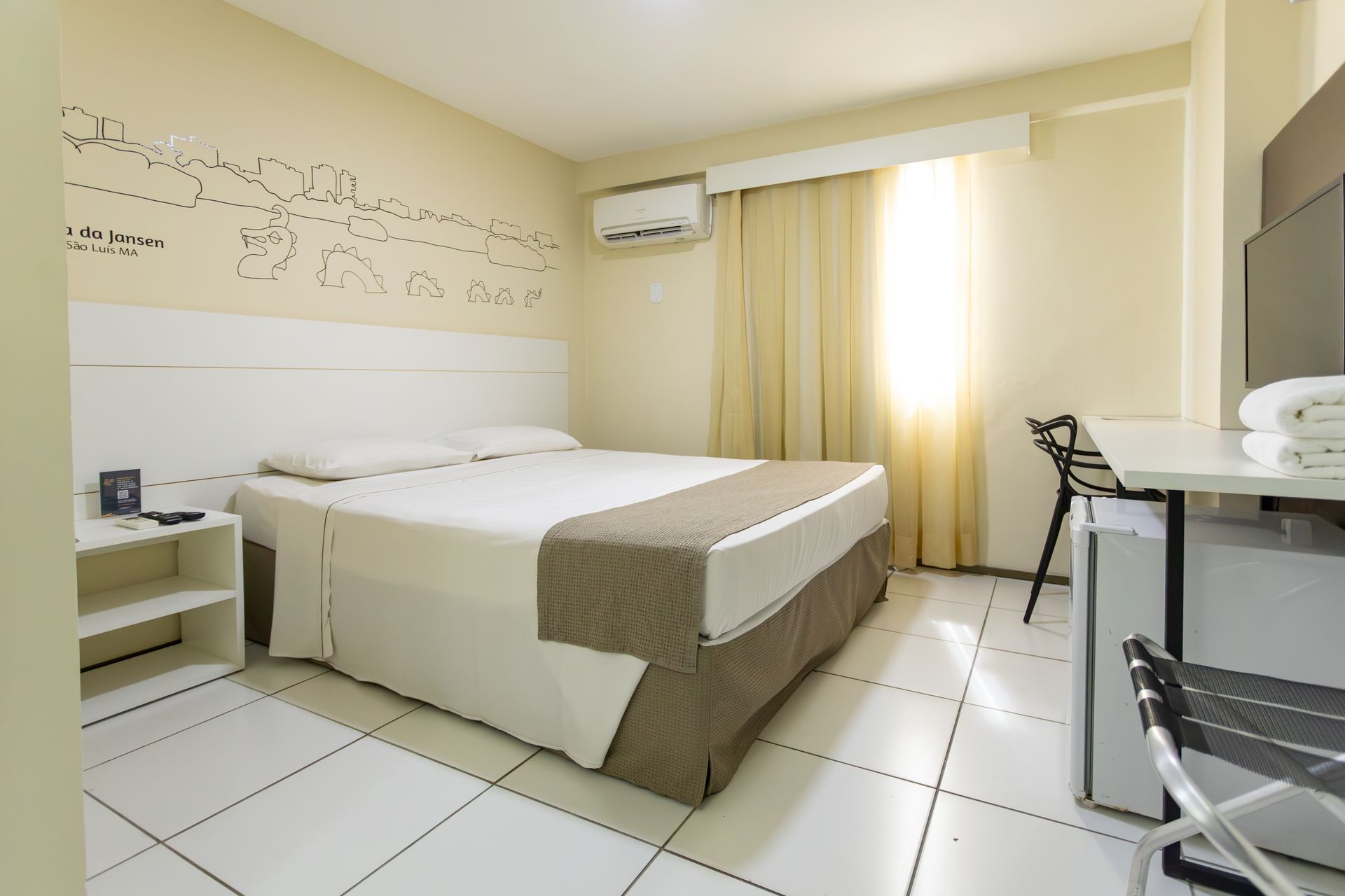 Quarto de hotel com cama de casal, escrivaninha pequena e televisão. Paredes brancas e piso de azulejo.