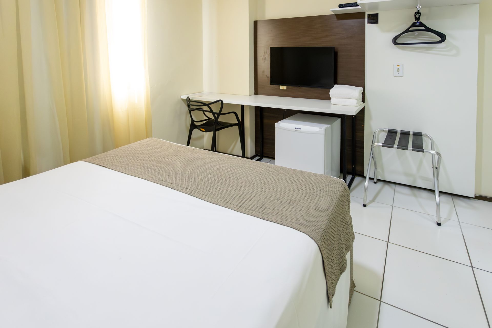 Quarto de hotel com cama, escrivaninha, TV, frigobar e suporte para bagagem.