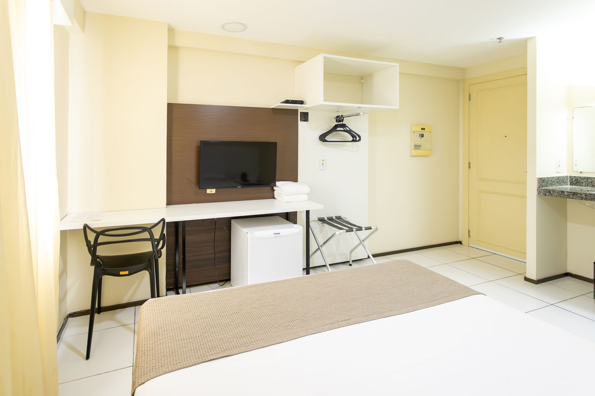 Quarto de hotel com cama, escrivaninha, TV, frigobar e porta.