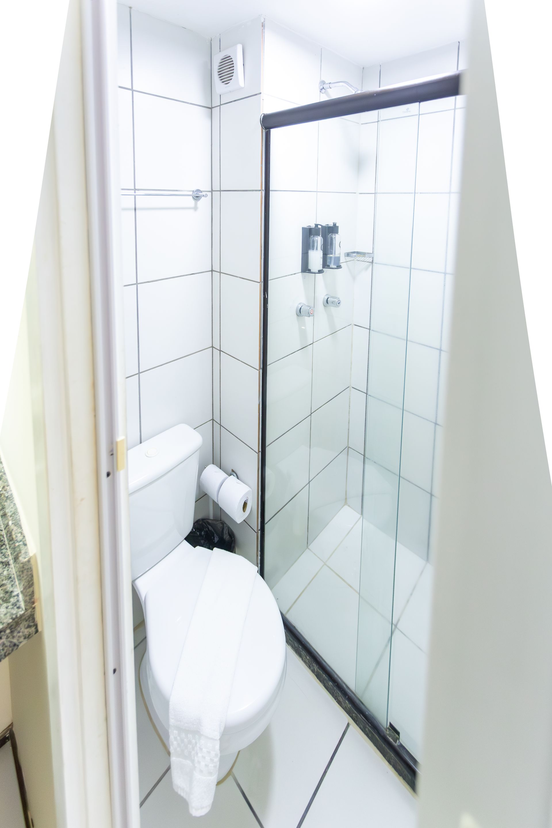 Pequeno banheiro branco com vaso sanitário, chuveiro e paredes revestidas com azulejos.