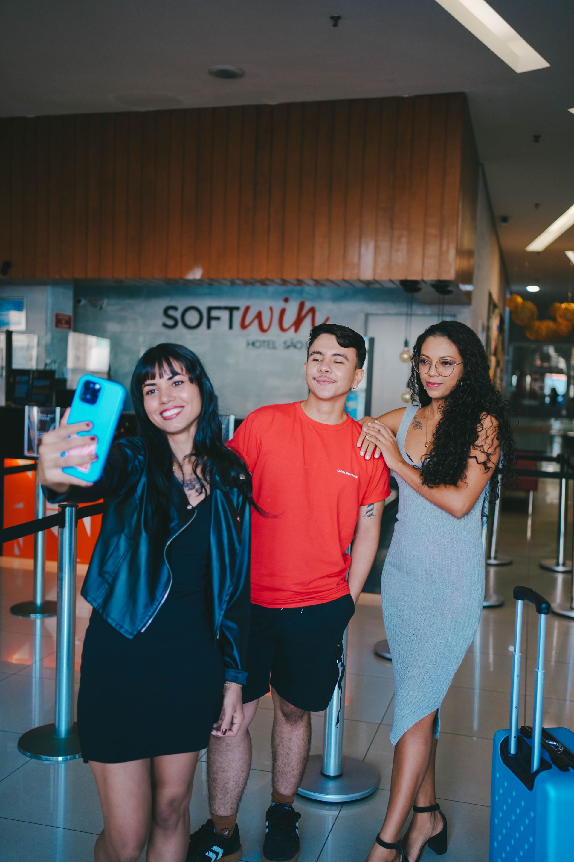 Três pessoas posam para uma selfie em frente à placa de um hotel. Uma delas segura o celular, todas sorrindo.